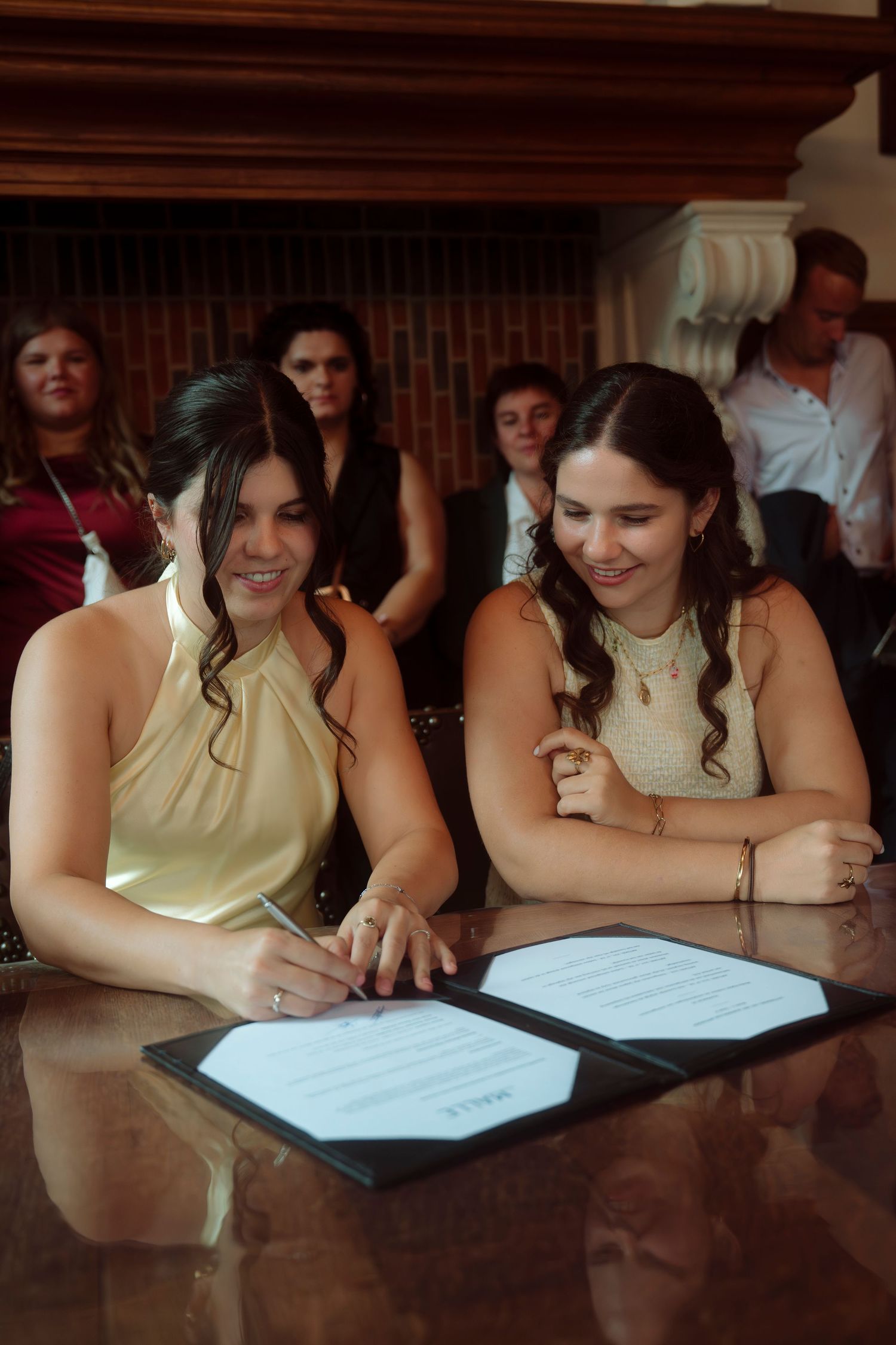 Two women smiling while signing a document at a table, surrounded by guests at a formal ceremony.