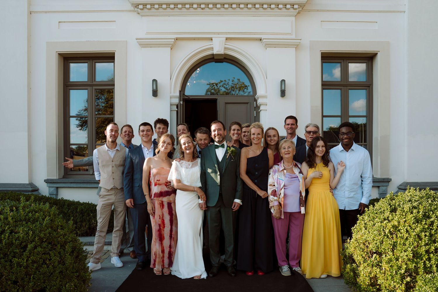 Wedding party group photo outside elegant white building with bride in white gown and groom in green suit.