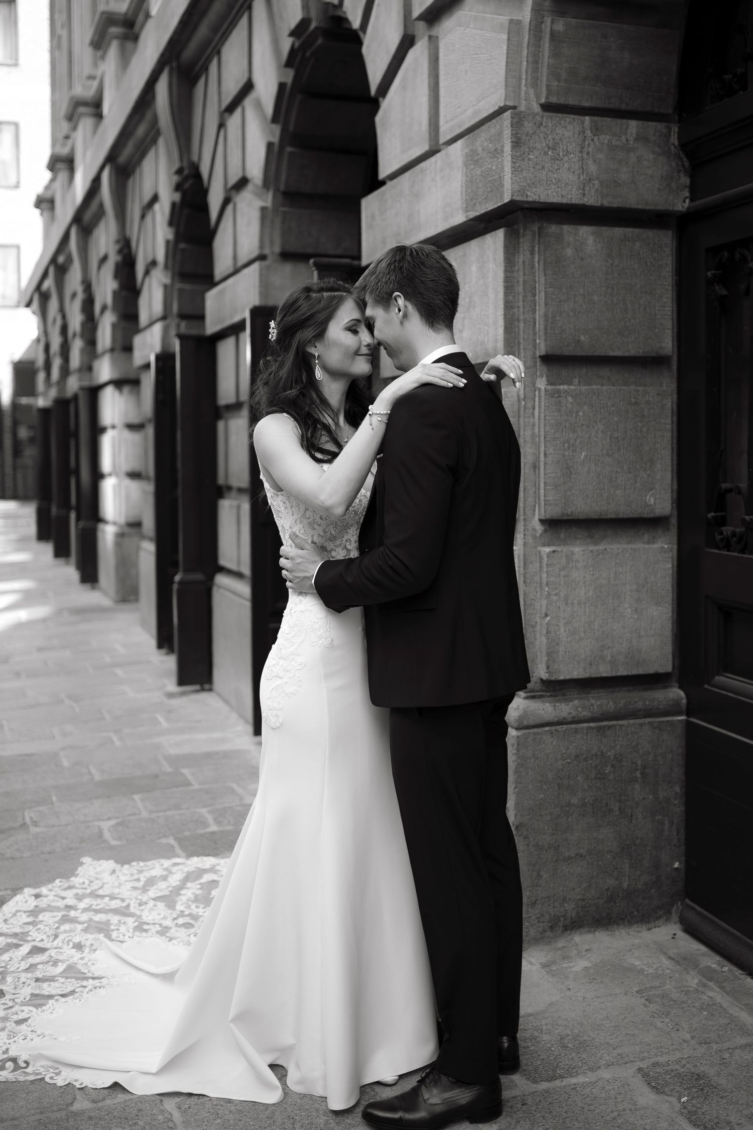 Black and white photo of a bride and groom embracing tenderly outside a classic stone building on their wedding day.