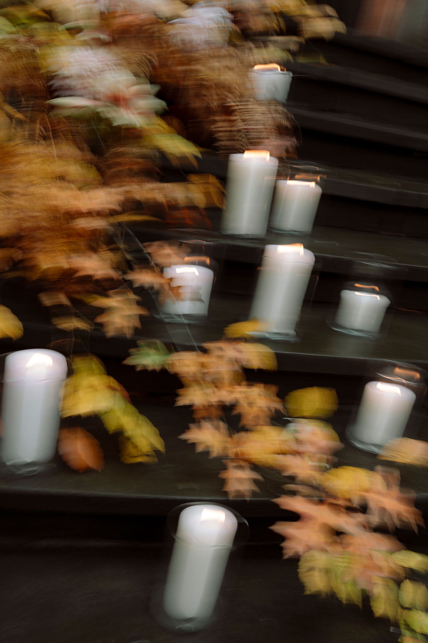 Blurred white candles glowing on dark steps surrounded by colorful autumn leaves in a moody, artistic long-exposure photo.