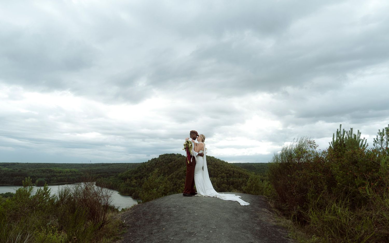 Bride and groom kissing on a rocky overlook with a dramatic cloudy sky and river landscape.