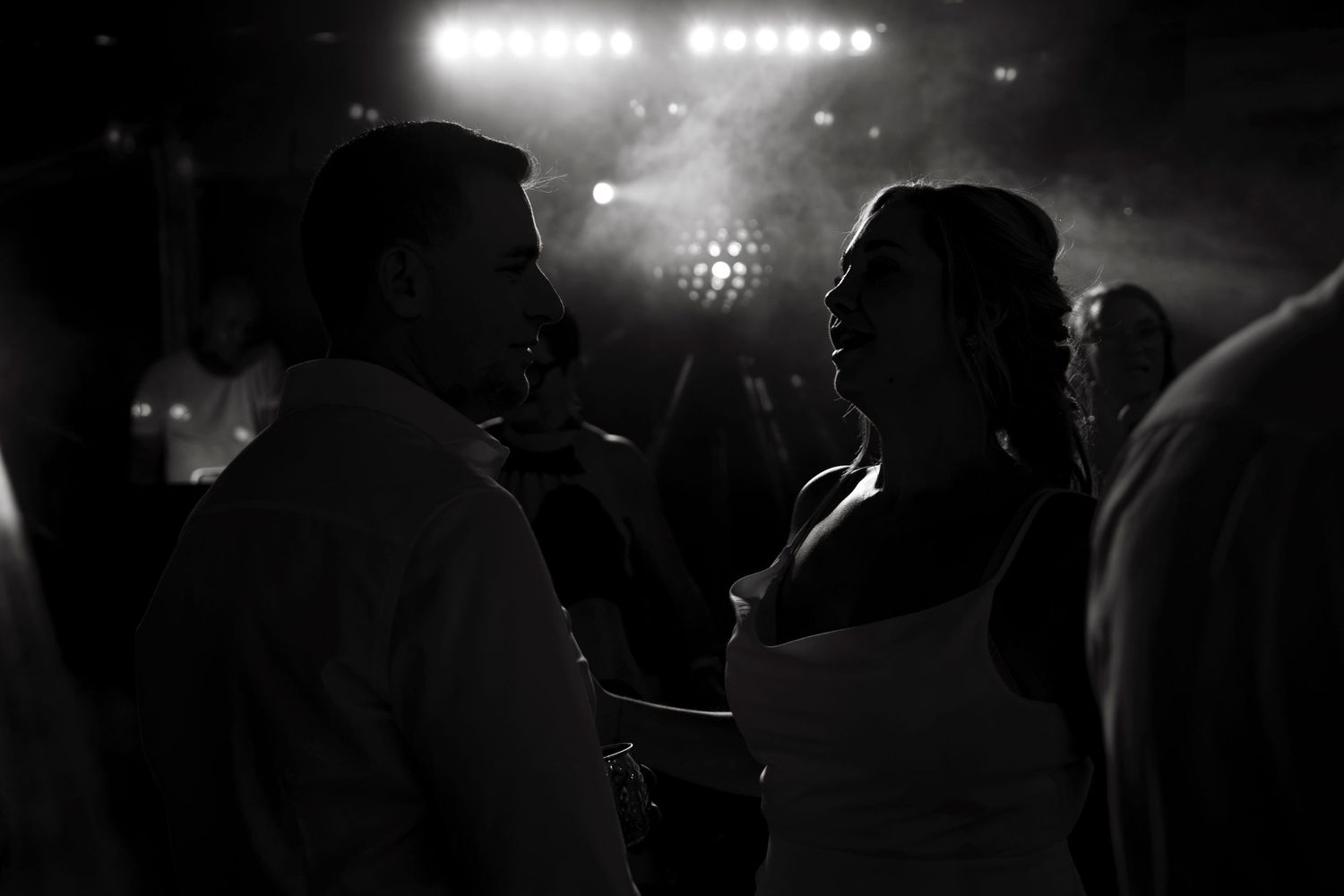 Black and white photo of a couple sharing a romantic dance at a wedding reception with stage lights glowing behind them.