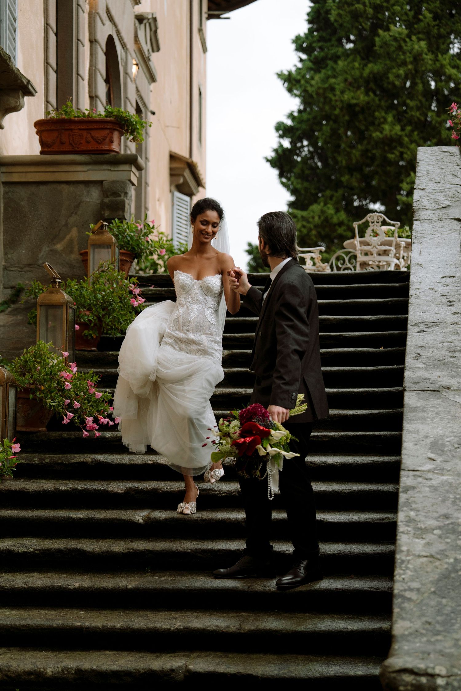 Bride and groom walk up stone steps at an Italian villa, bride holding her white gown and a burgundy bouquet.