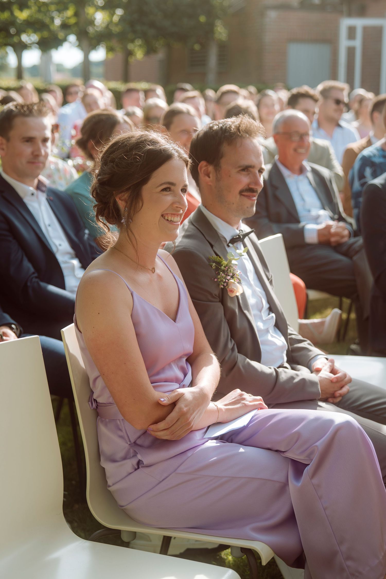 Smiling woman in lavender dress seated at outdoor wedding ceremony with guests in background.