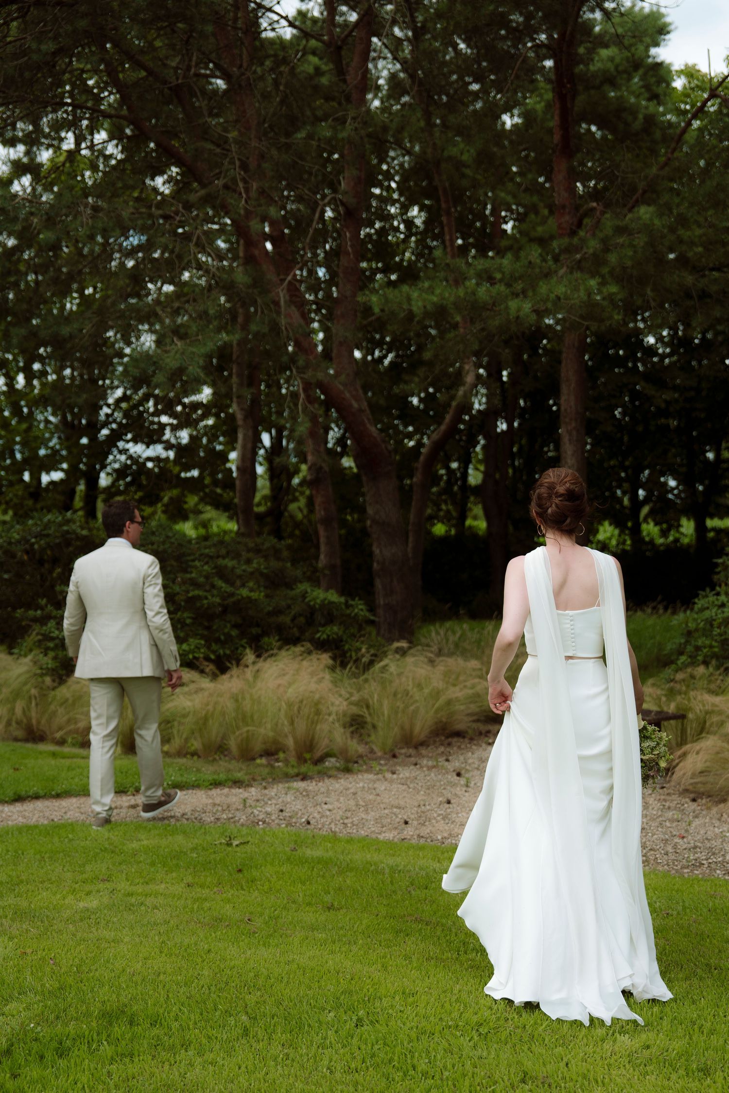 Bride in white gown and groom in tan suit walk toward trees in a lush green garden wedding photo.