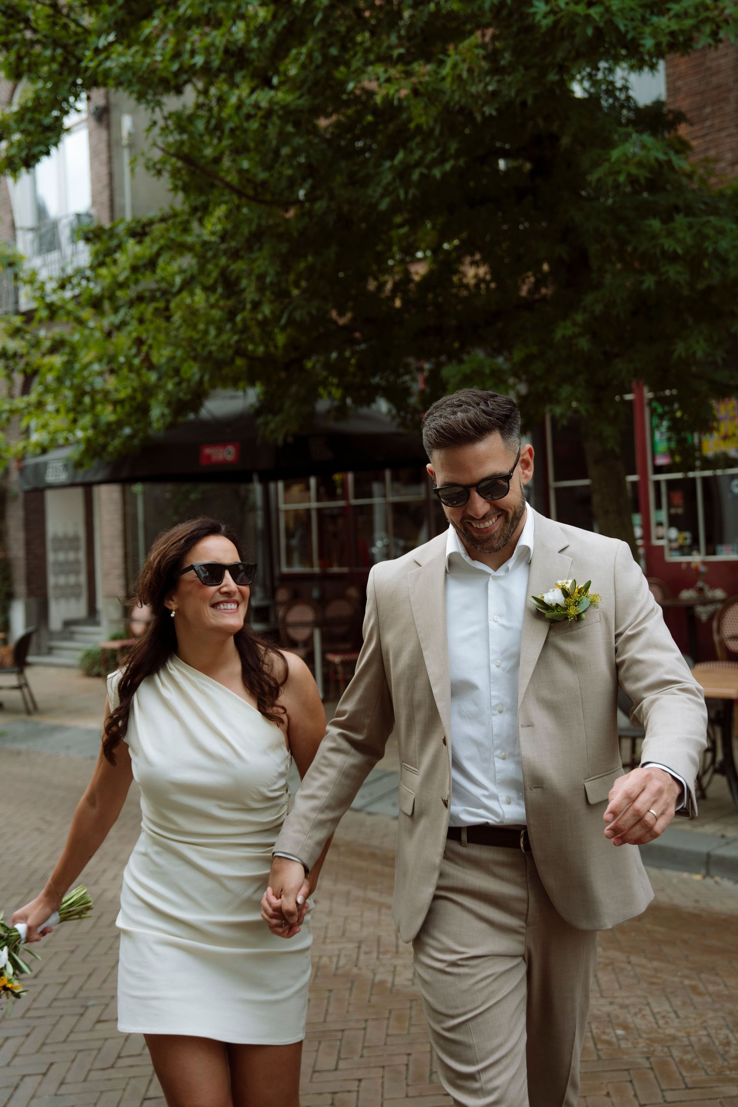 Stylish couple in sunglasses holding hands and smiling while walking on city street on wedding day.