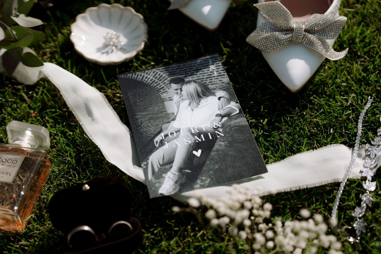 Black and white wedding photo card placed on green grass surrounded by rings, flowers, and elegant accessories.