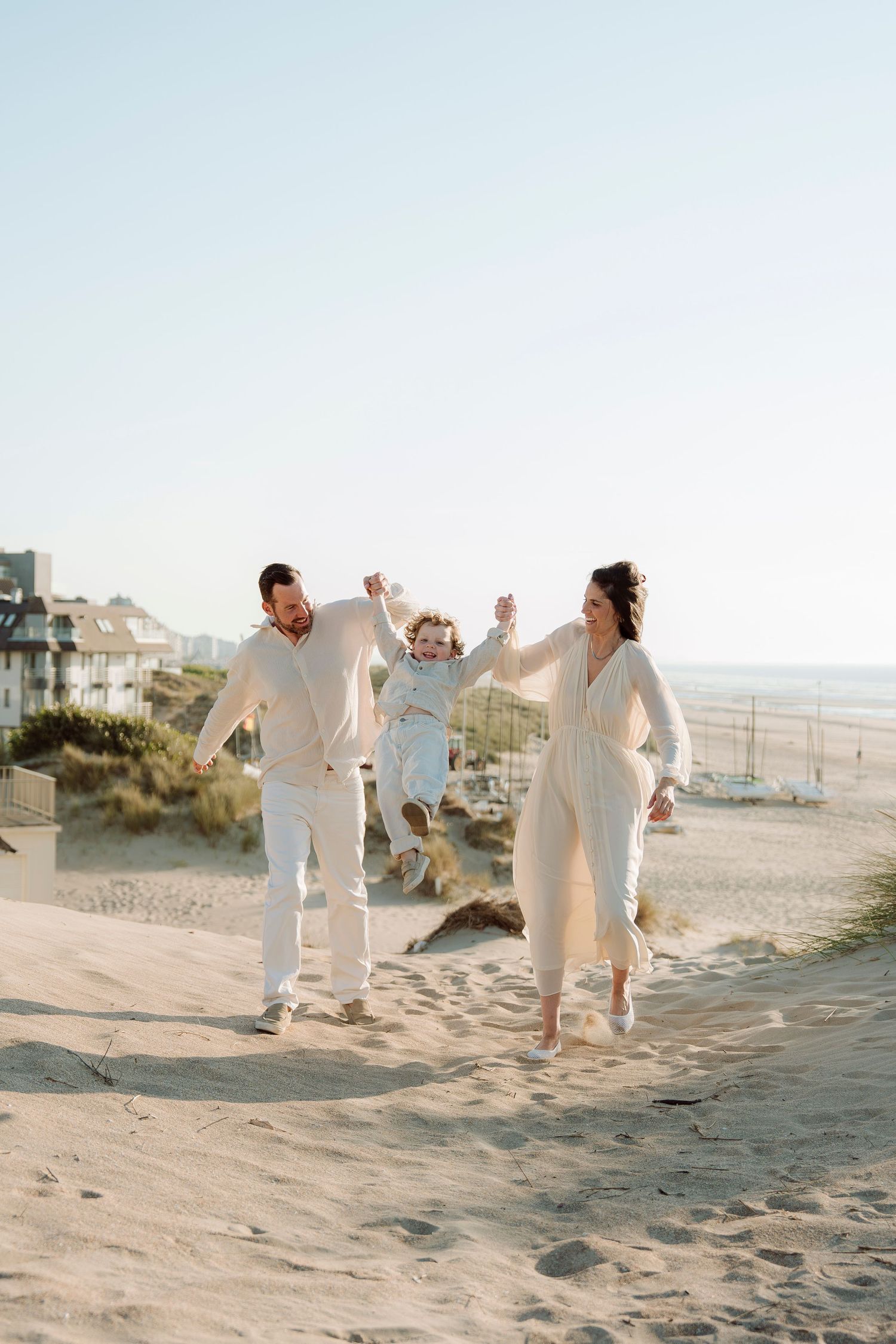 A joyful family swings their young child on a sunny beach, all dressed in white, with ocean and buildings in the background.