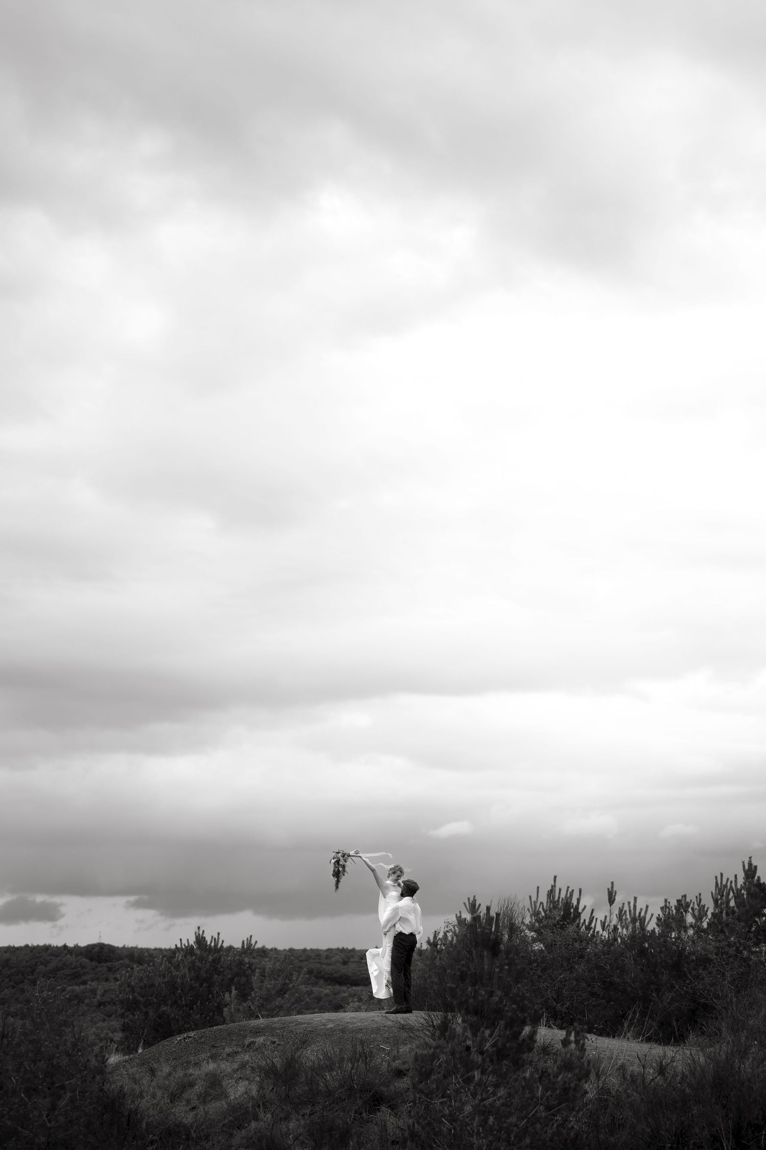 Black and white photo of a couple embracing on a rocky hilltop, raising a bouquet under dramatic cloudy skies.