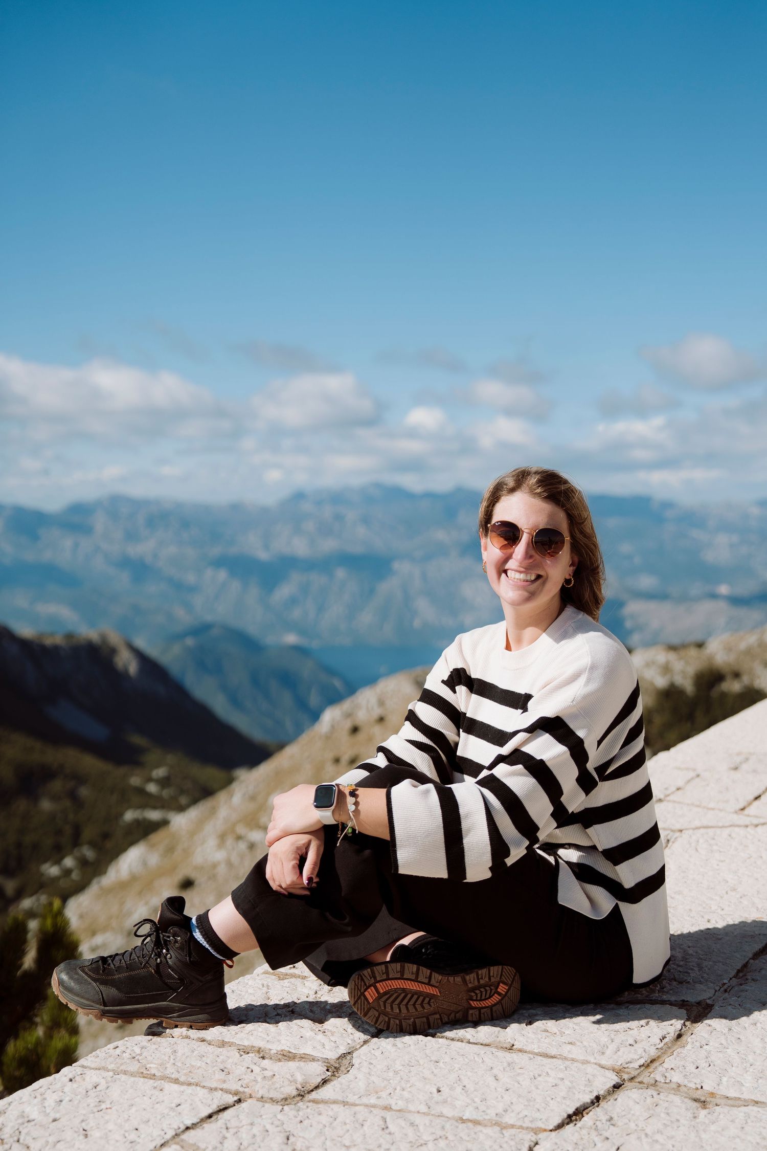 Smiling woman in striped sweater and sunglasses sitting on mountain viewpoint with scenic alpine landscape.