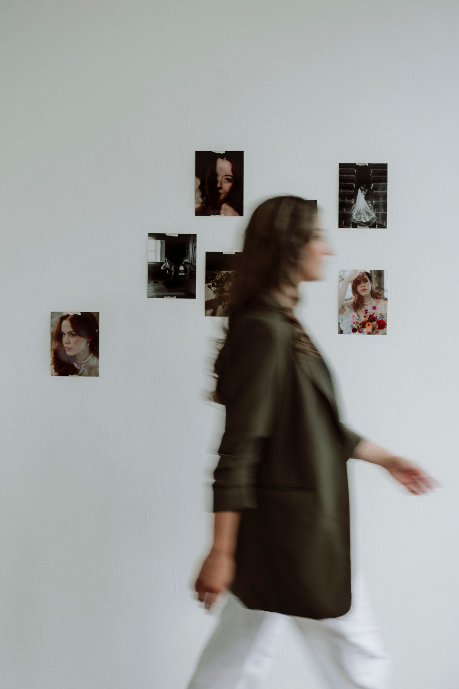 Blurred woman in dark jacket walking past white wall with photo collage in modern gallery space.