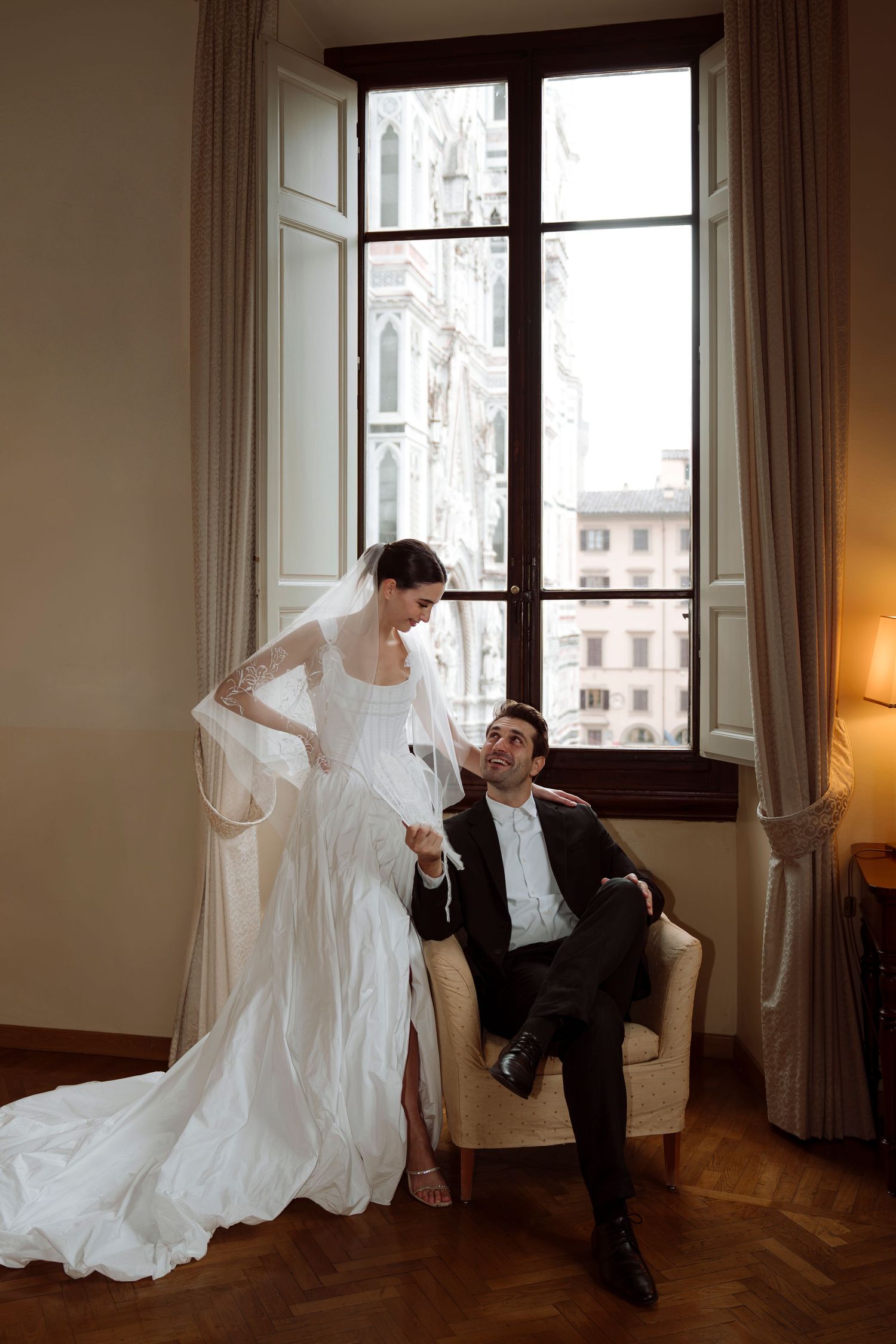 Elegant bride in white gown and veil stands by groom seated in armchair, with Florence cathedral view through tall window.