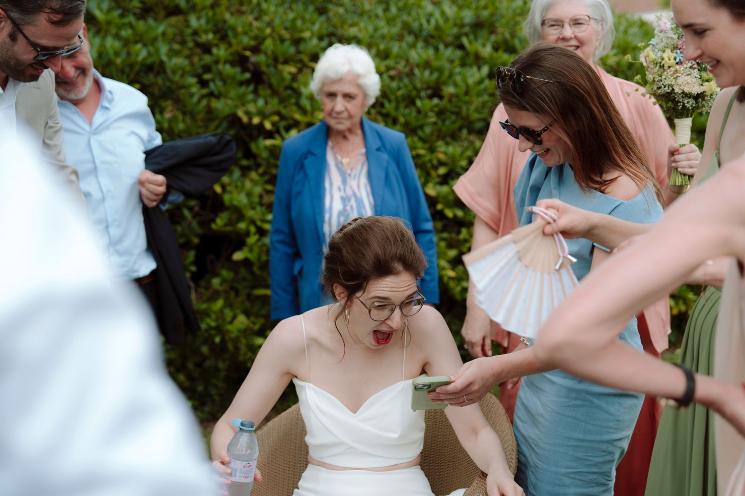 Bride in white dress reading a card surrounded by smiling guests and bridesmaids at an outdoor wedding reception.