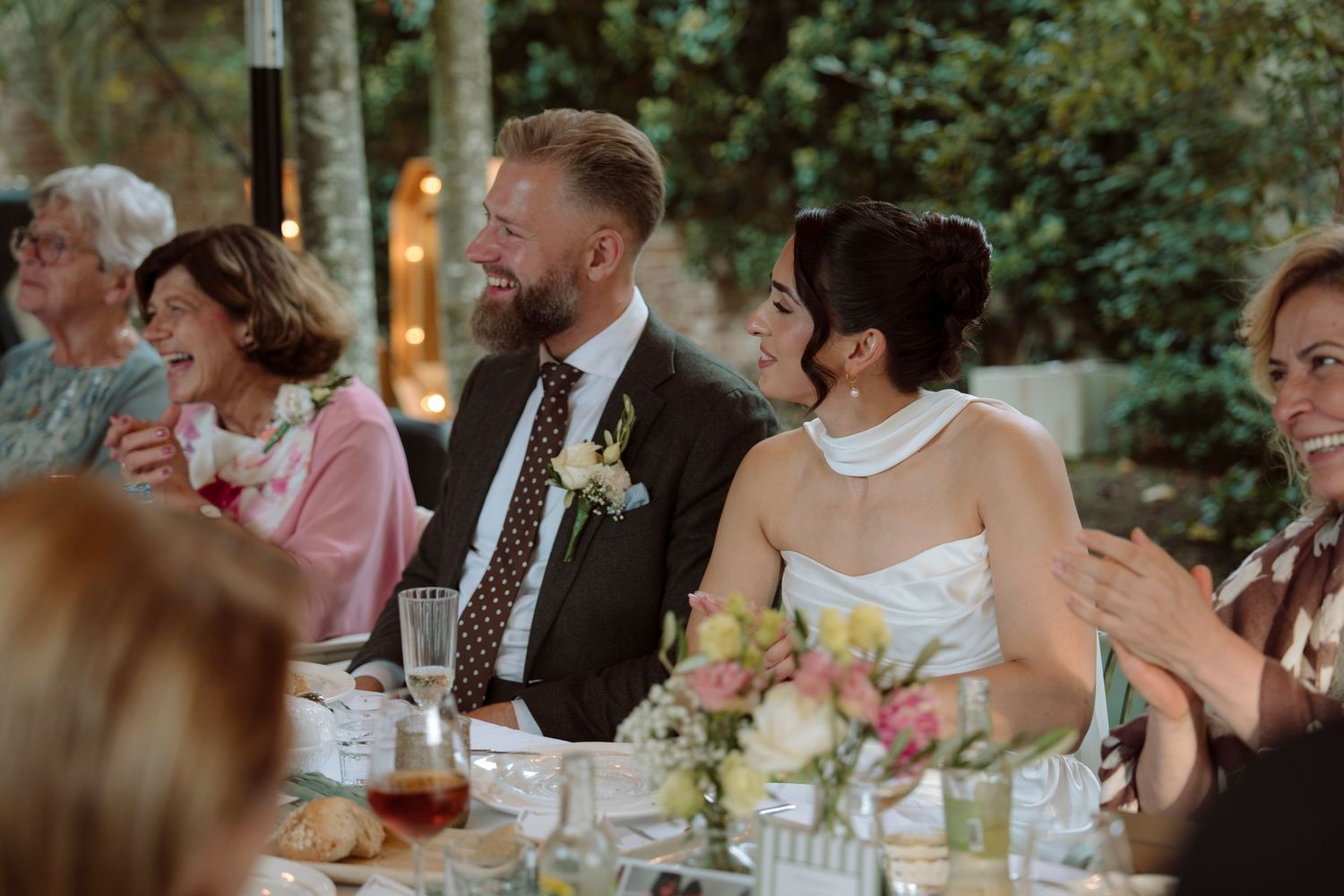 A smiling bride and groom sit together at their outdoor wedding reception dinner table surrounded by guests.