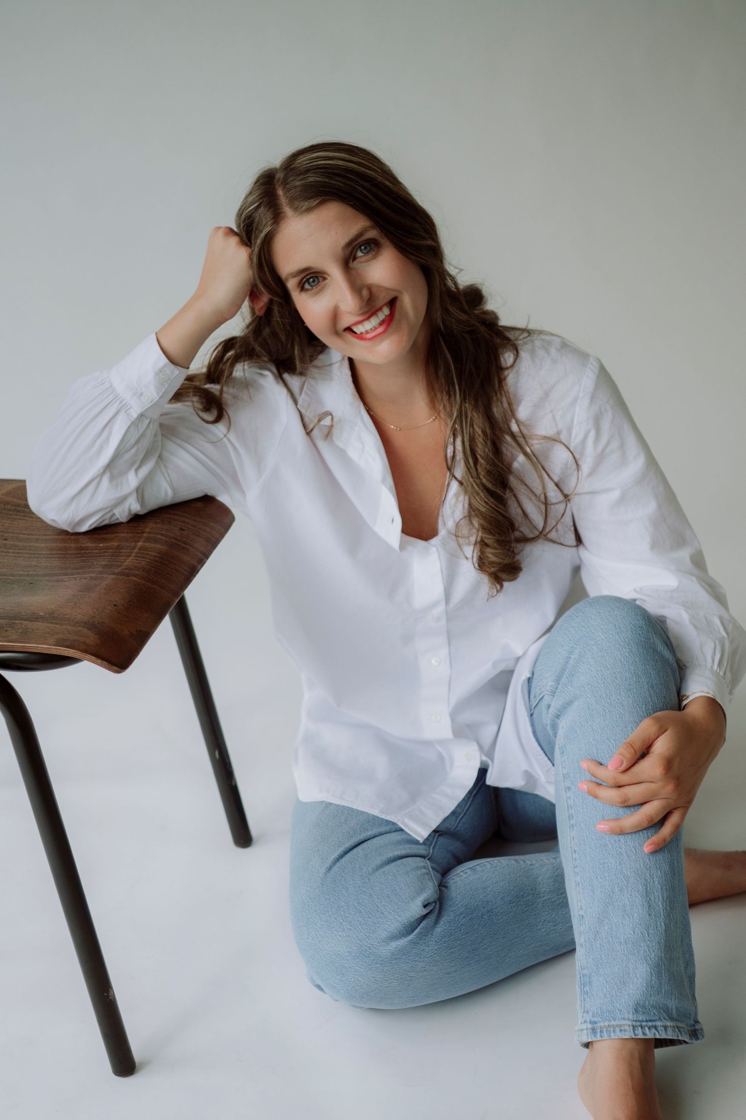 Smiling woman in white shirt and jeans sitting on floor beside wooden chair in bright studio.