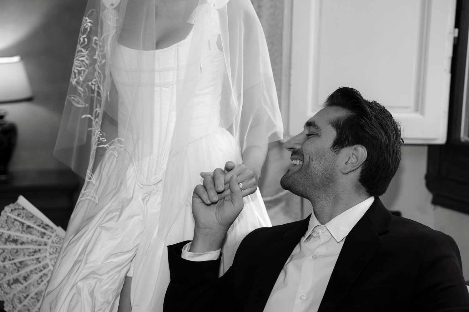 Black and white photo of a groom laughing joyfully while kneeling before a bride in a white wedding dress.
