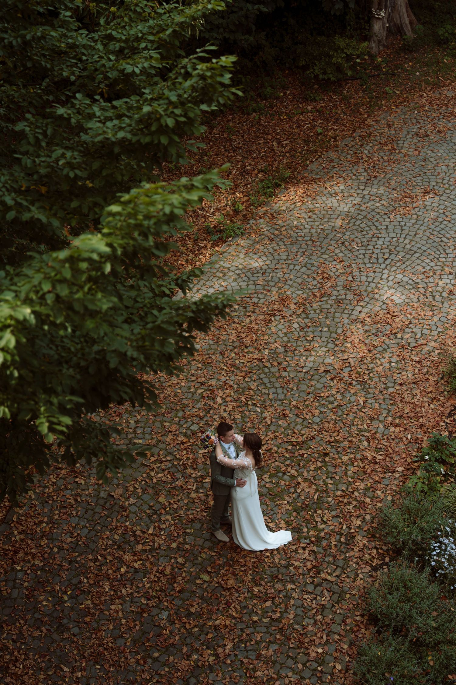 Aerial view of bride and groom embracing on cobblestone path surrounded by autumn leaves and lush green trees.