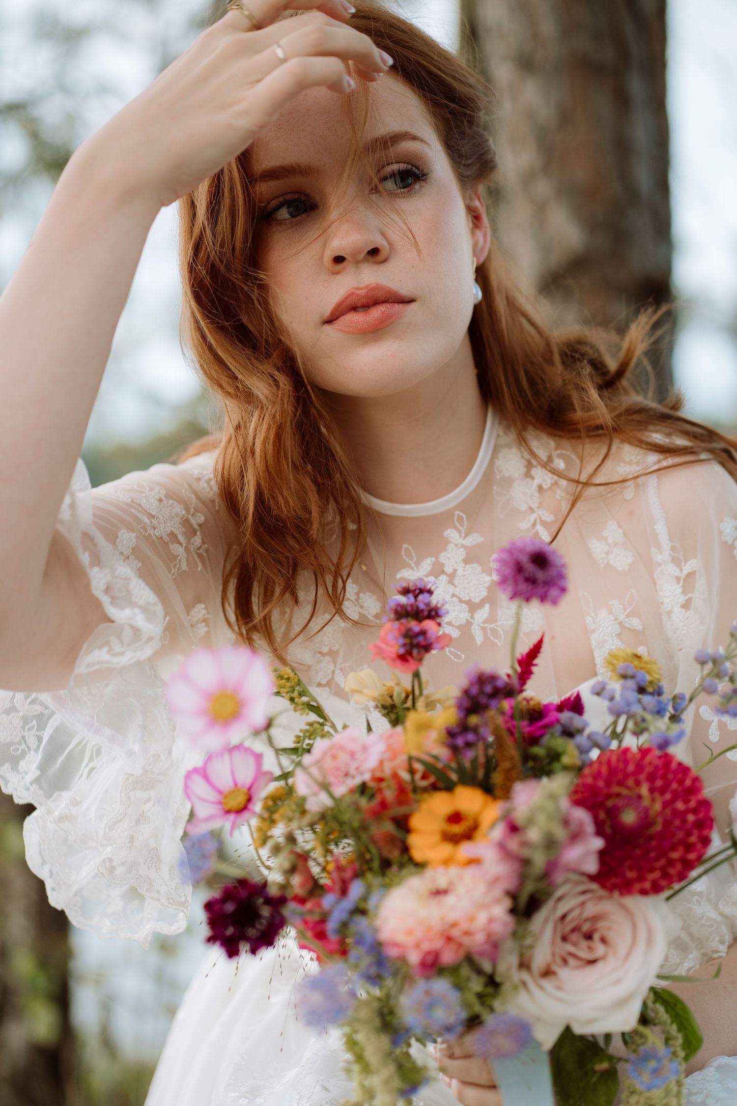 Bride in lace dress holding colorful wildflower bouquet, posing outdoors near trees with hand raised to forehead.