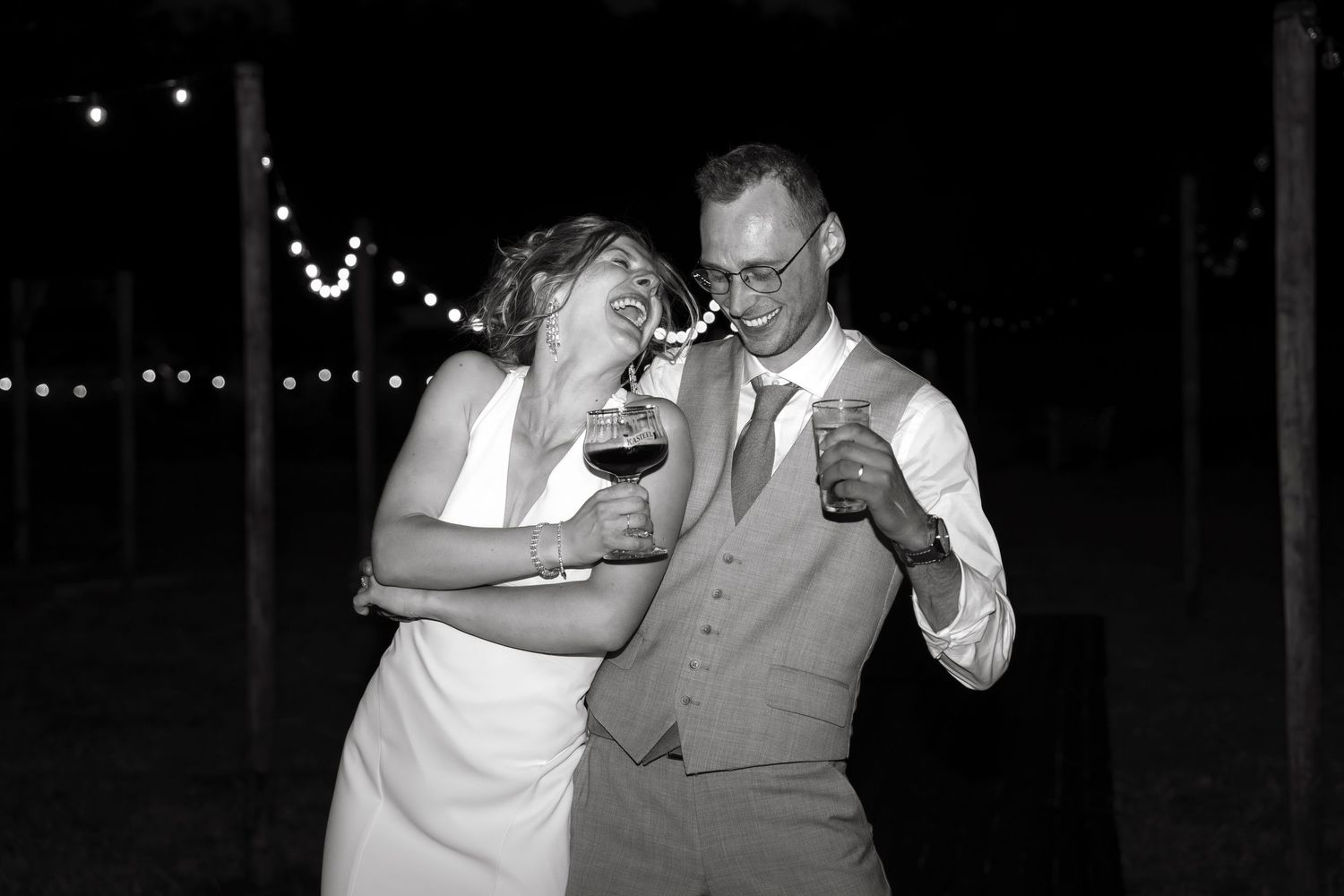 Black and white photo of a laughing bride and groom holding drinks at an outdoor wedding reception with string lights.