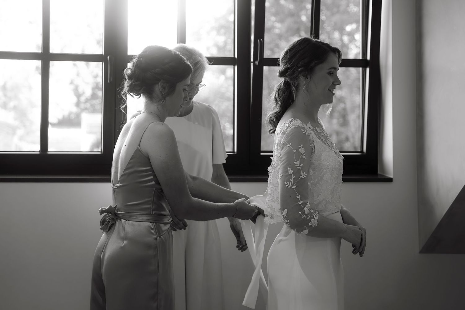 Black and white photo of bride smiling as bridesmaids help her prepare near sunlit windows on wedding day.