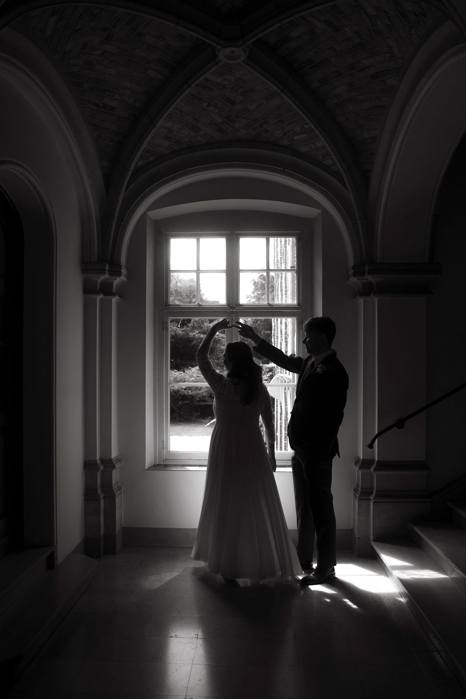 Black and white photo of bride and groom dancing in elegant arched hallway with sunlight streaming through window.