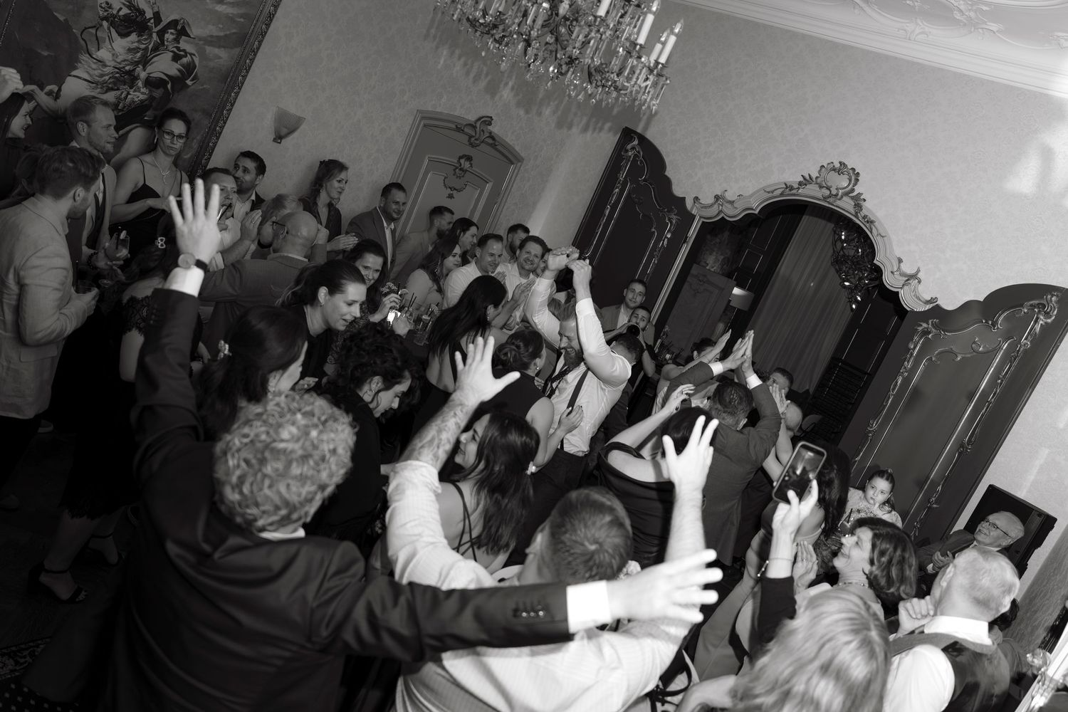 Black and white photo of a lively wedding reception crowd with guests dancing and raising hands in elegant ballroom.