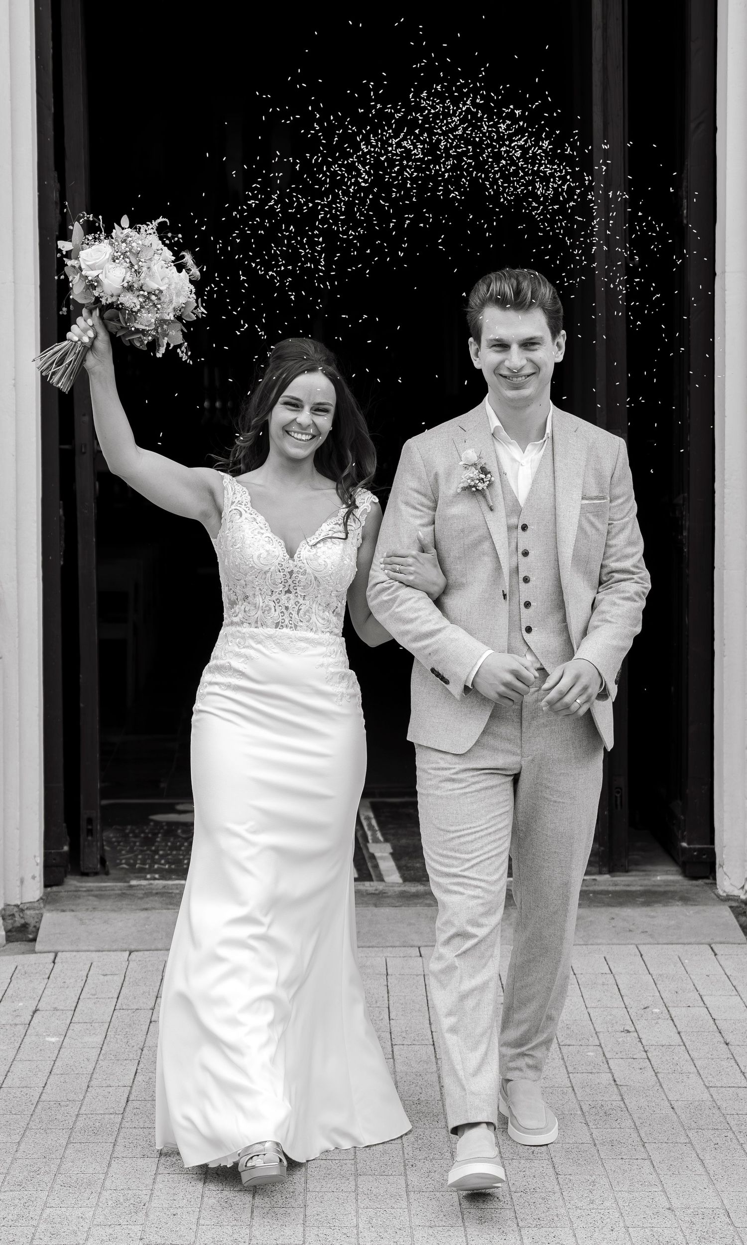 Black and white photo of a smiling bride holding bouquet aloft and groom in suit exiting church after wedding ceremony.
