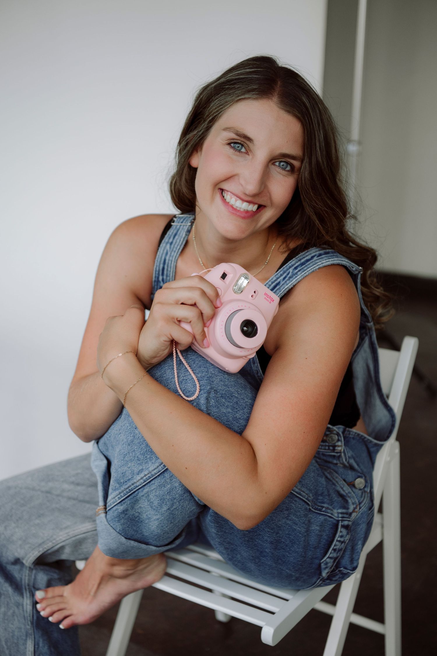 Smiling woman in denim overalls holding a pink Fujifilm Instax Mini camera while sitting on a white chair.