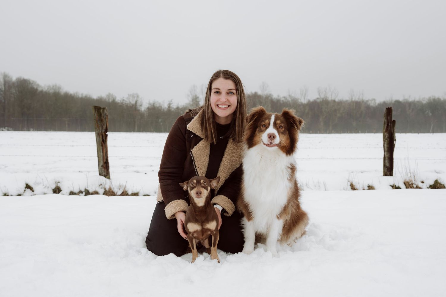 Young woman kneeling in snow with two dogs, a Chihuahua and Australian Shepherd, in a snowy winter field.