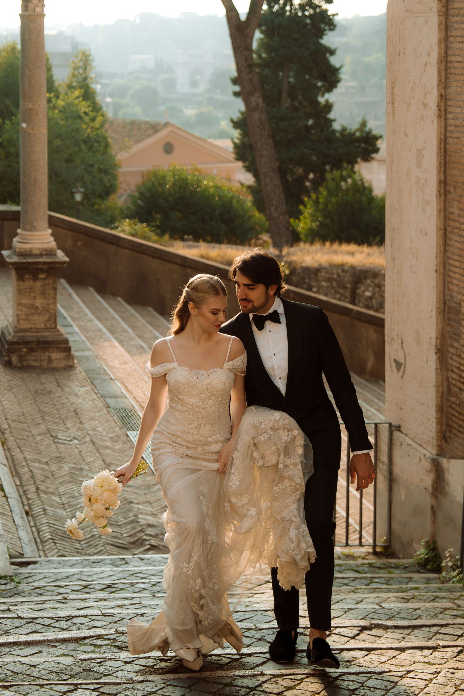 Bride and groom walk cobblestone steps in Rome, she in lace gown with bouquet, he in black tuxedo, golden sunset behind them.
