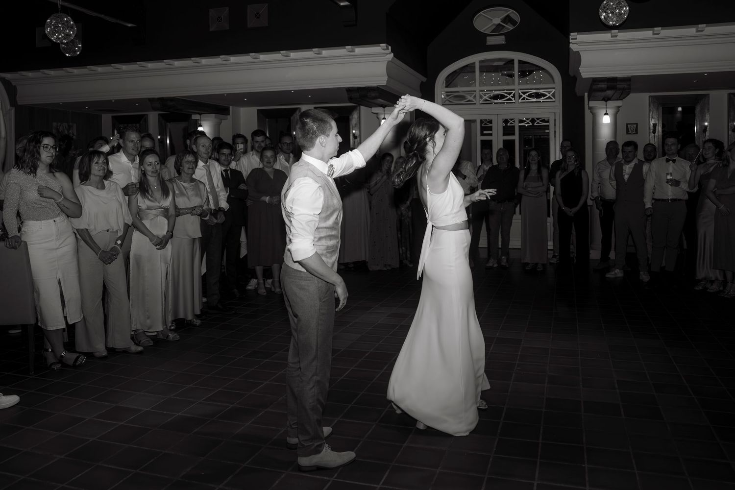 Black and white photo of bride and groom sharing first dance at wedding reception with guests watching.