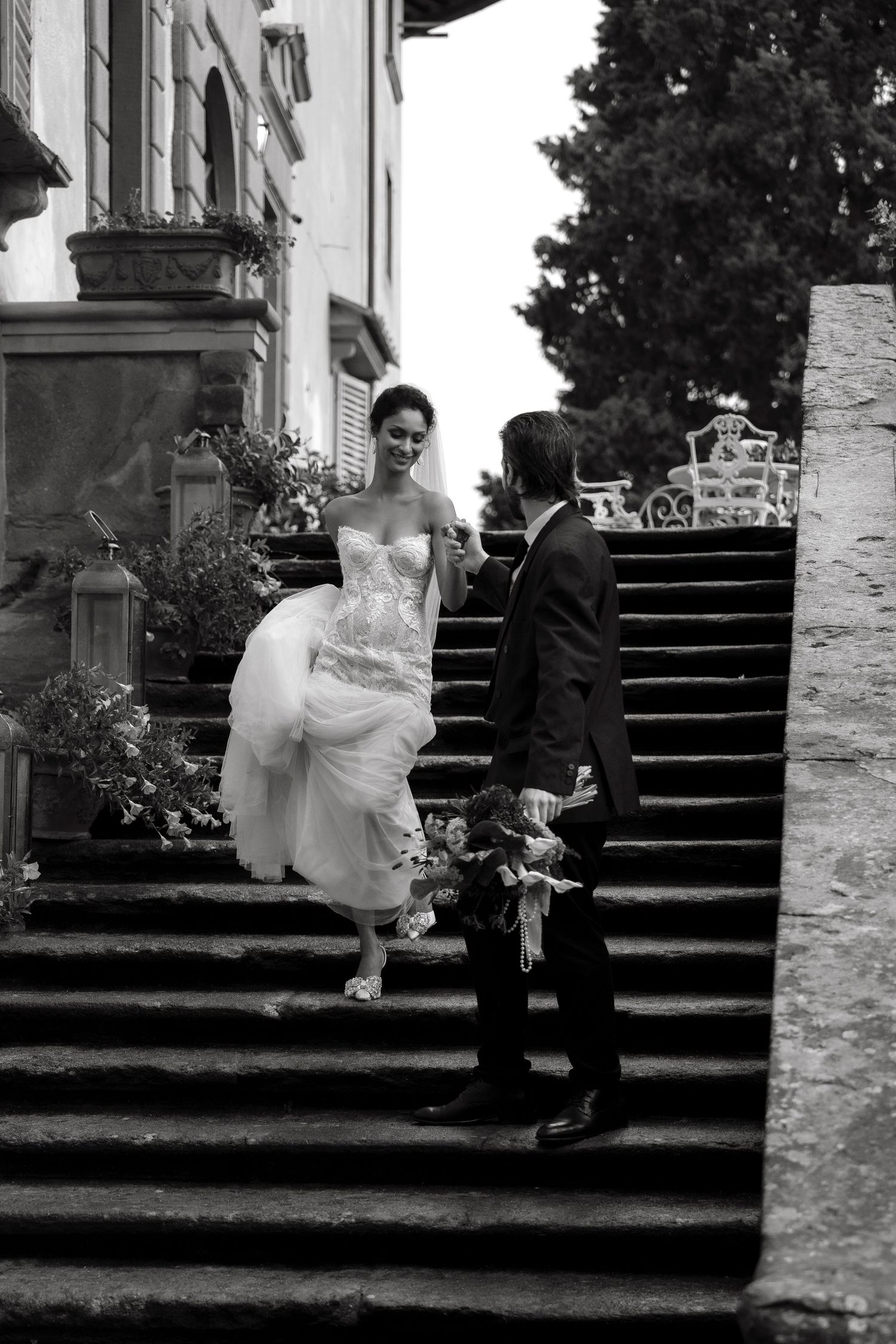 Black and white photo of a bride and groom on stone steps outside a villa, she holds her dress while he carries a bouquet.