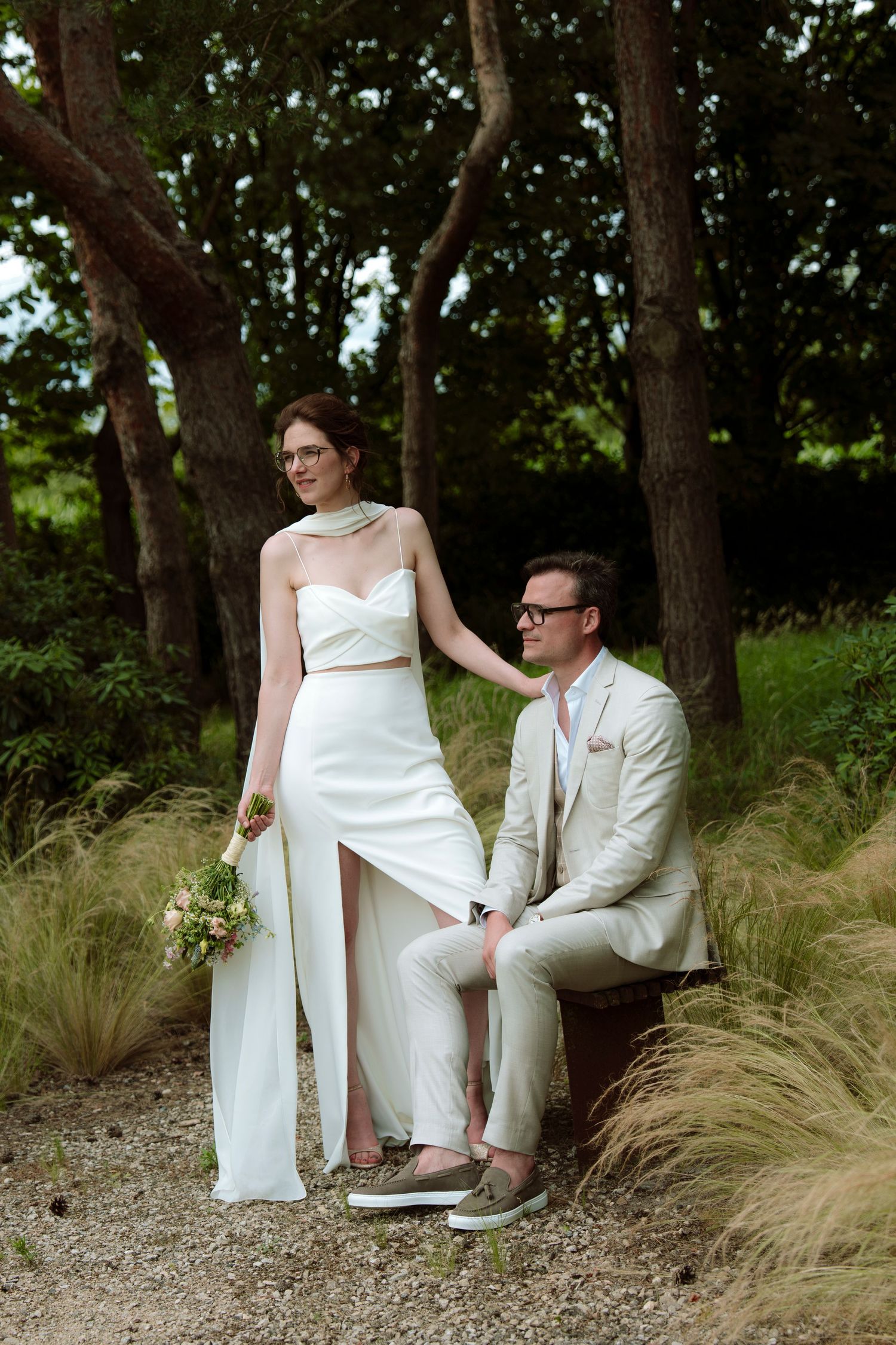 Bride in white two-piece gown holds bouquet beside groom in beige suit seated on luggage, surrounded by lush greenery.