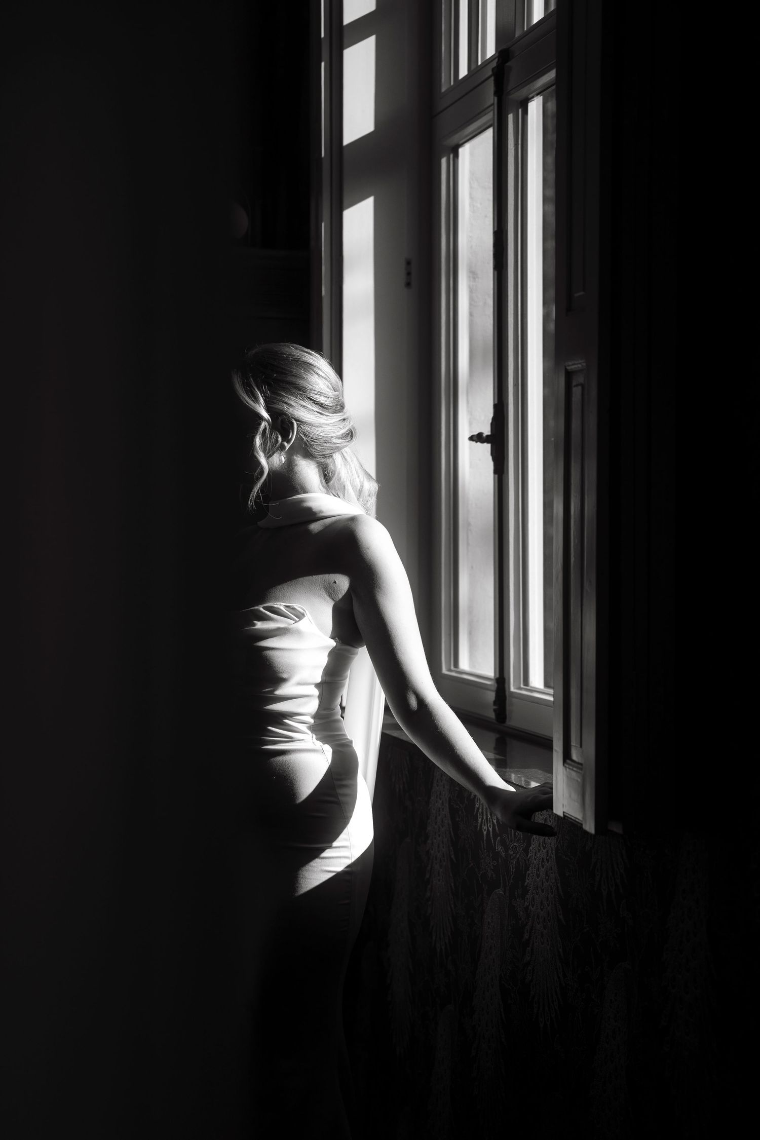 Black and white photo of a blonde woman in a white dress gazing out a sunlit window in a dark room.