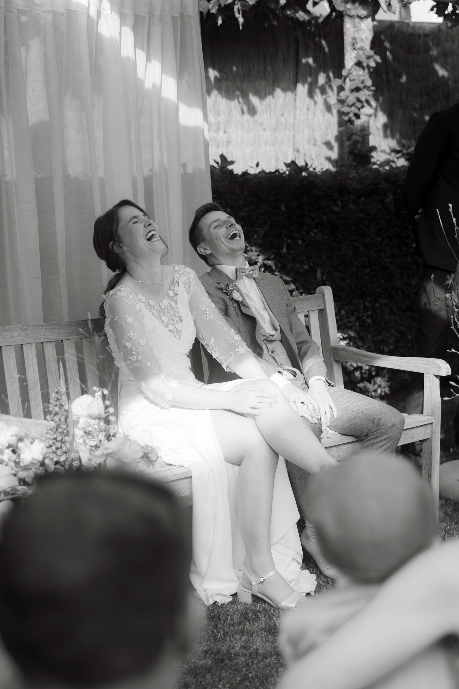 Black and white photo of a laughing bride and groom sitting on a bench during their outdoor wedding ceremony.