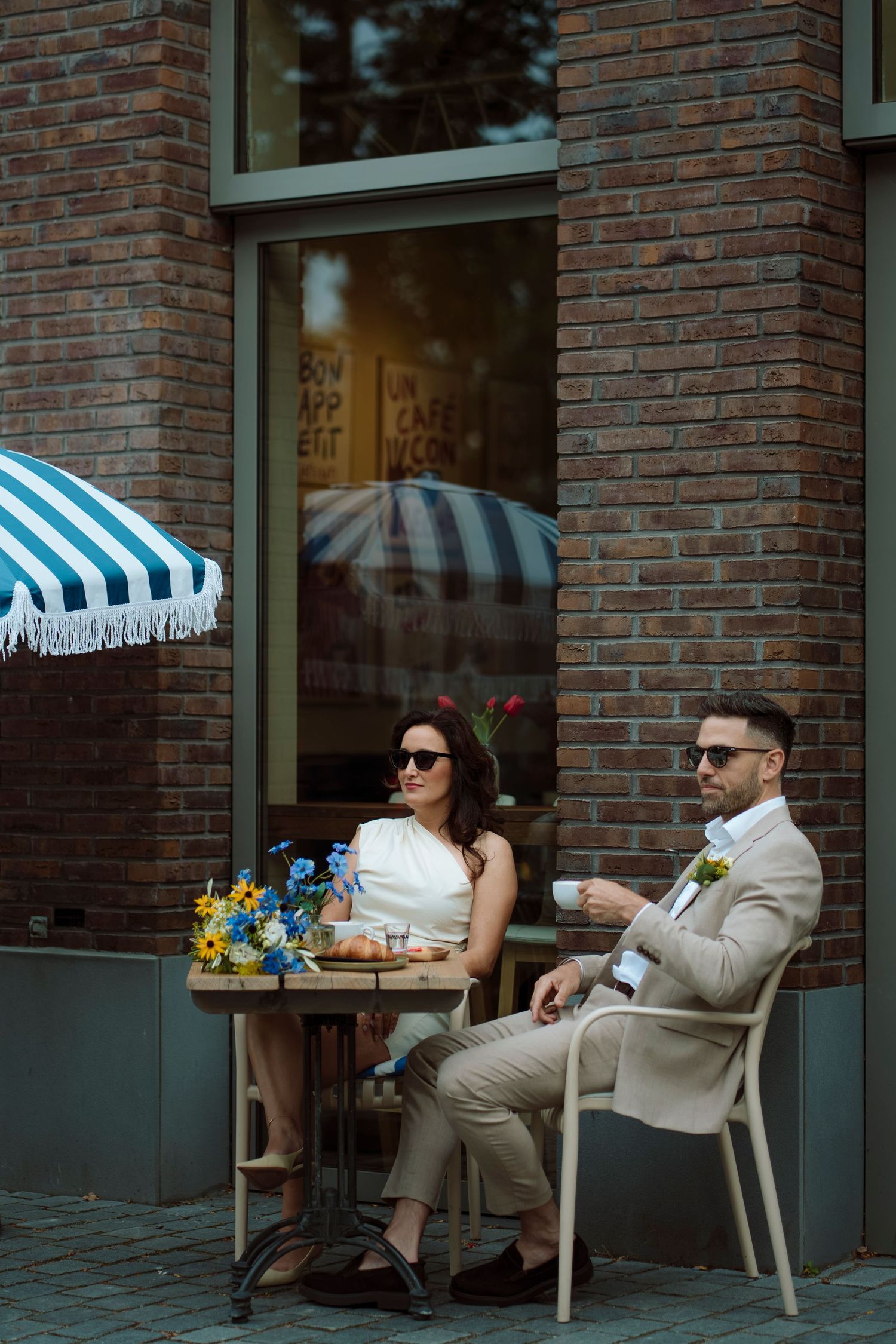 Elegant couple in sunglasses sitting at outdoor cafe table with flowers, man in beige suit holding coffee cup.