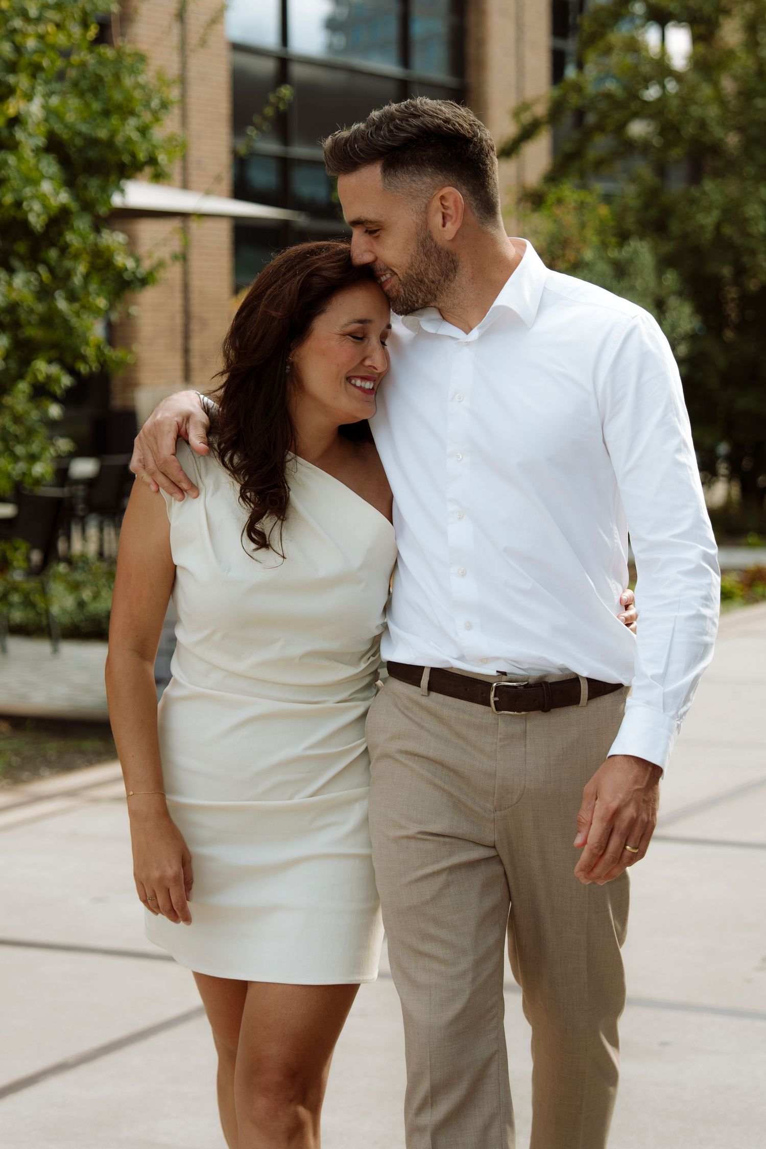 Smiling groom in white shirt embraces bride in white dress during outdoor wedding portrait.
