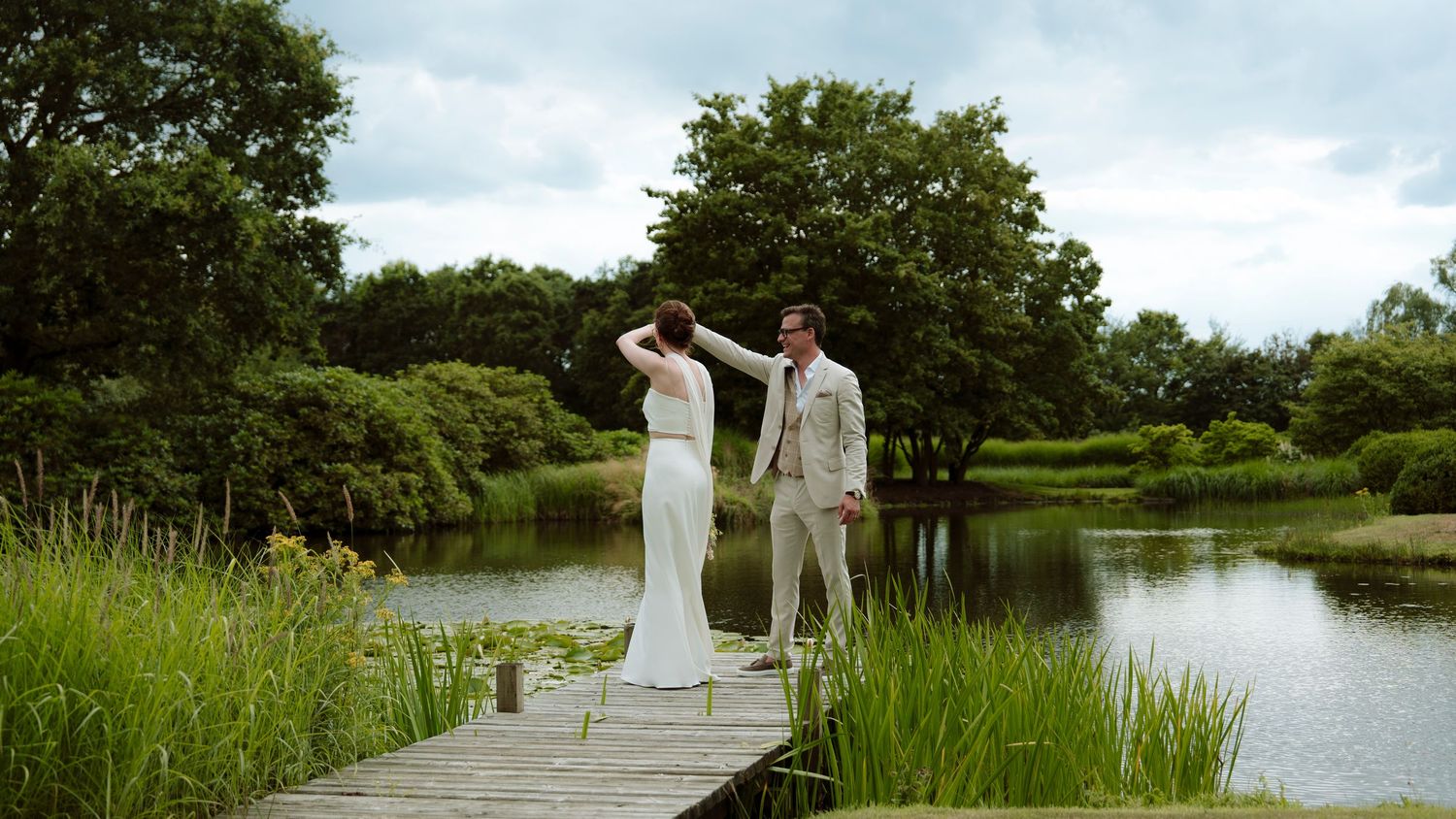 Bride and groom standing on a wooden dock by a serene pond surrounded by lush green trees and tall grass.