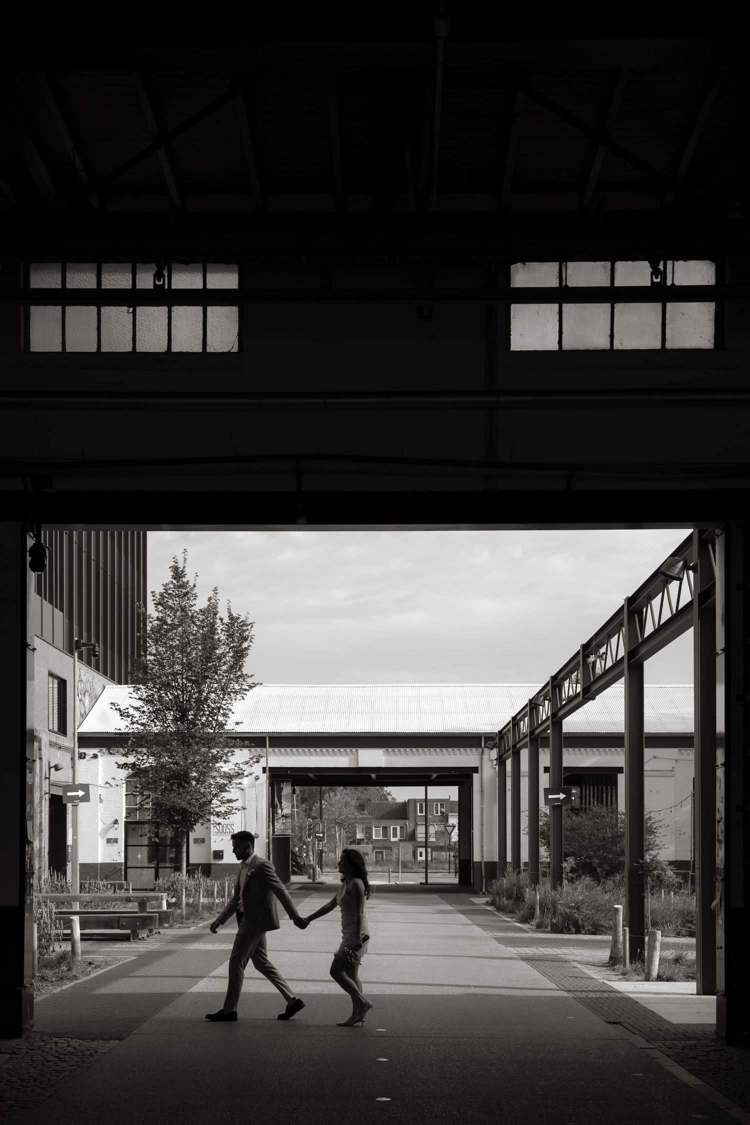Silhouette of bride and groom holding hands walking through industrial building archway in black and white.