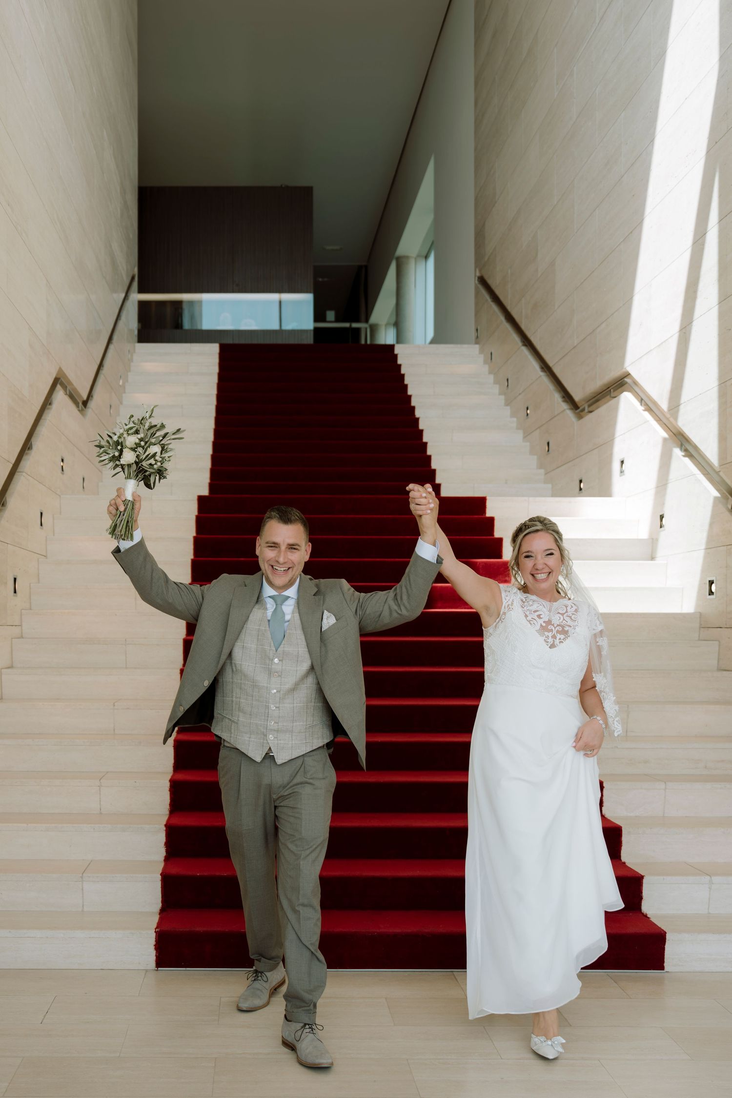 Bride and groom celebrate on red carpeted stairs, raising hands with bouquet in modern venue.
