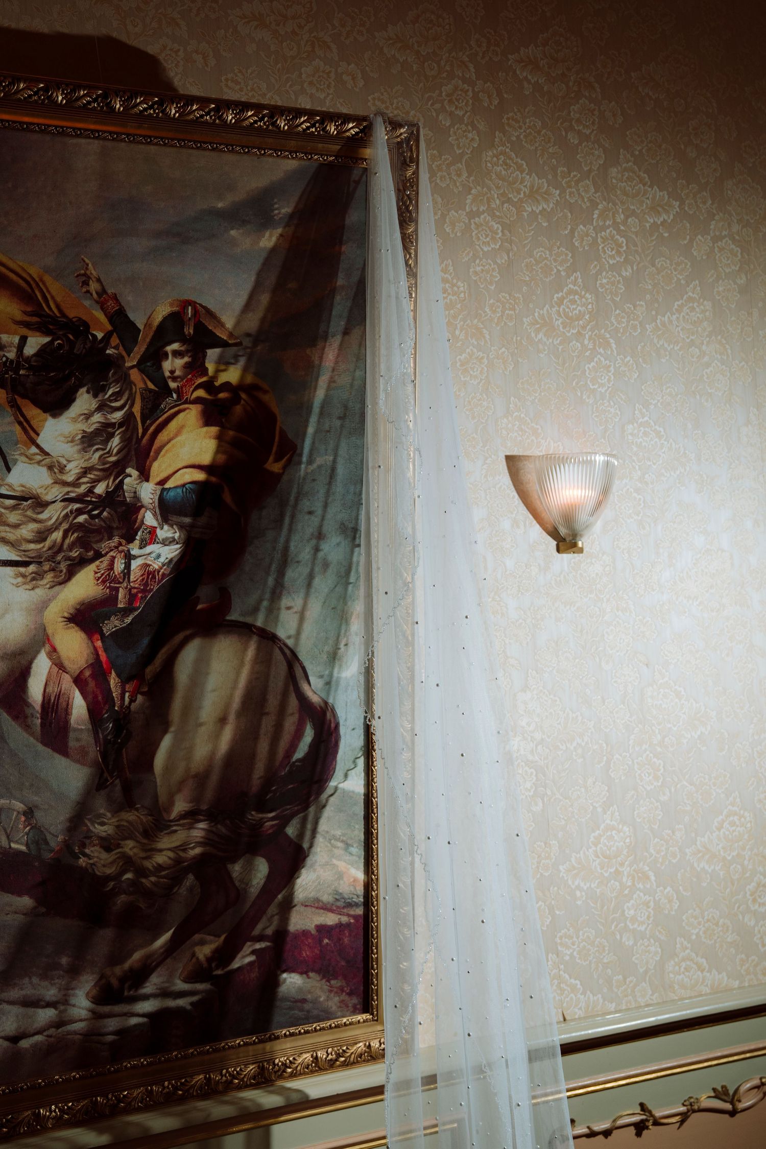 A bridal veil draped over a framed Napoleon painting on a damask wall with a vintage sconce light.