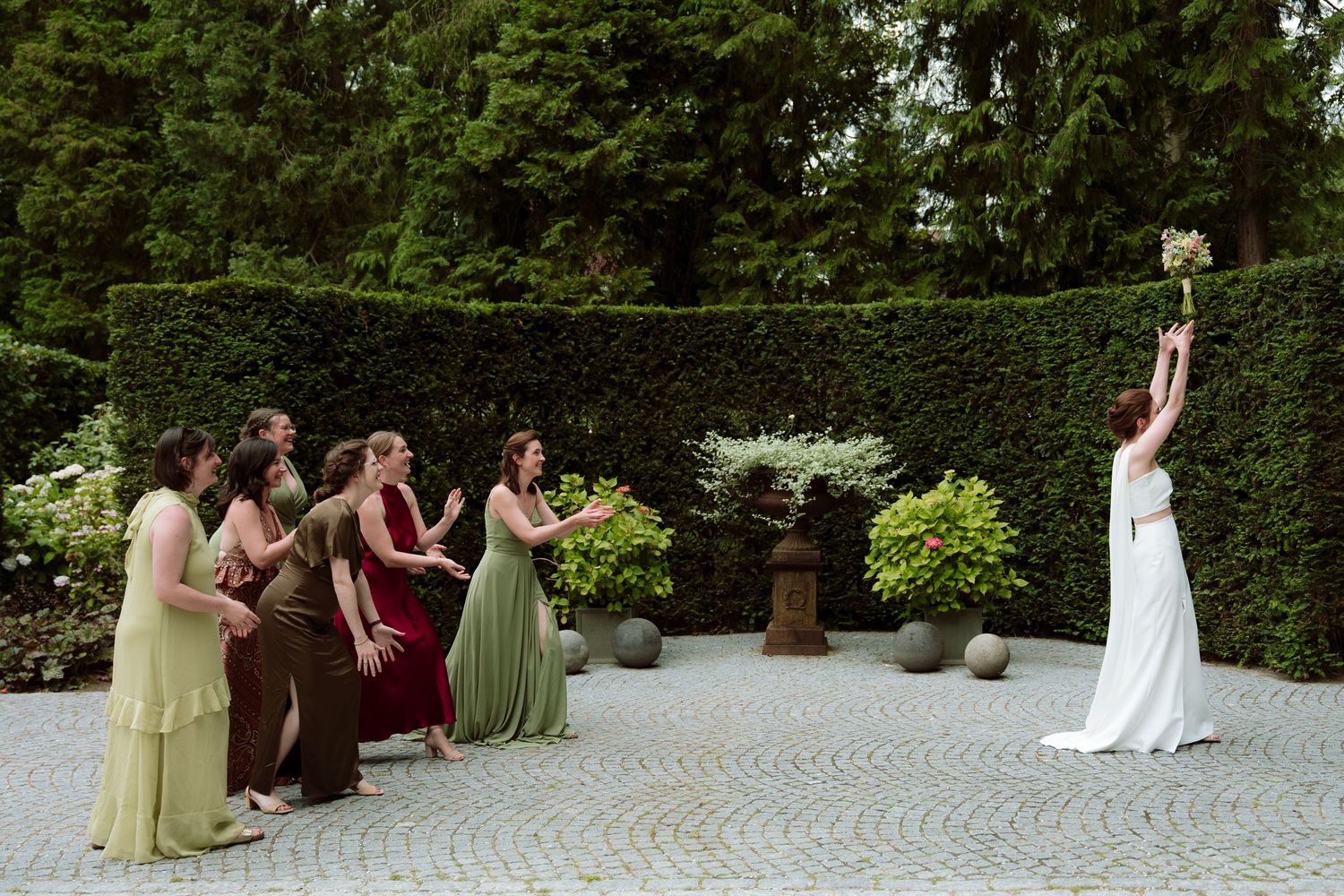 Bride tossing bouquet to bridesmaids in colorful dresses at outdoor garden wedding ceremony.