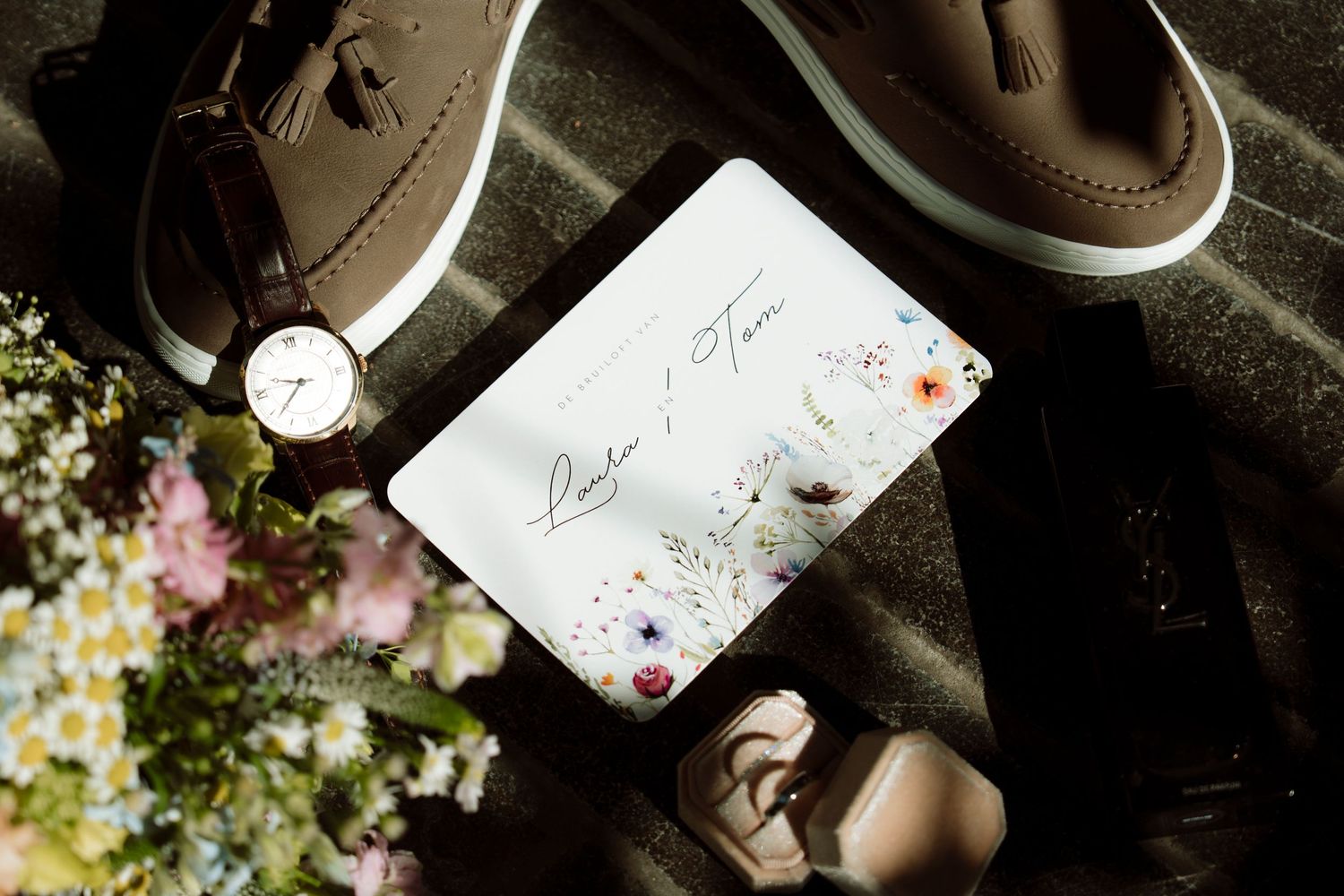 Flat lay of wedding invitation with floral design, brown loafers, watch, and flowers on dark surface.