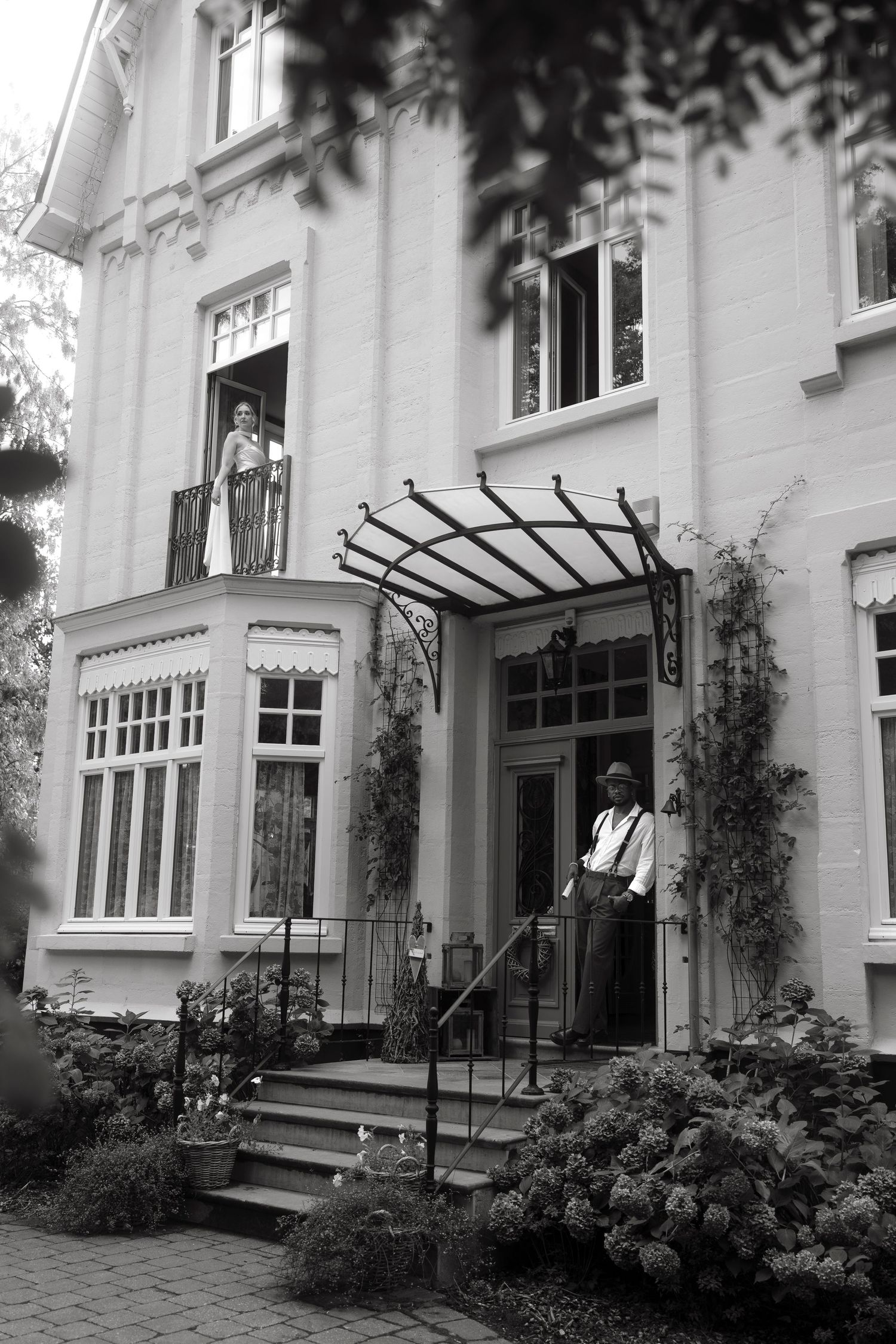 Black and white photo of a grand historic house with a man at the entrance and a woman on the balcony above.
