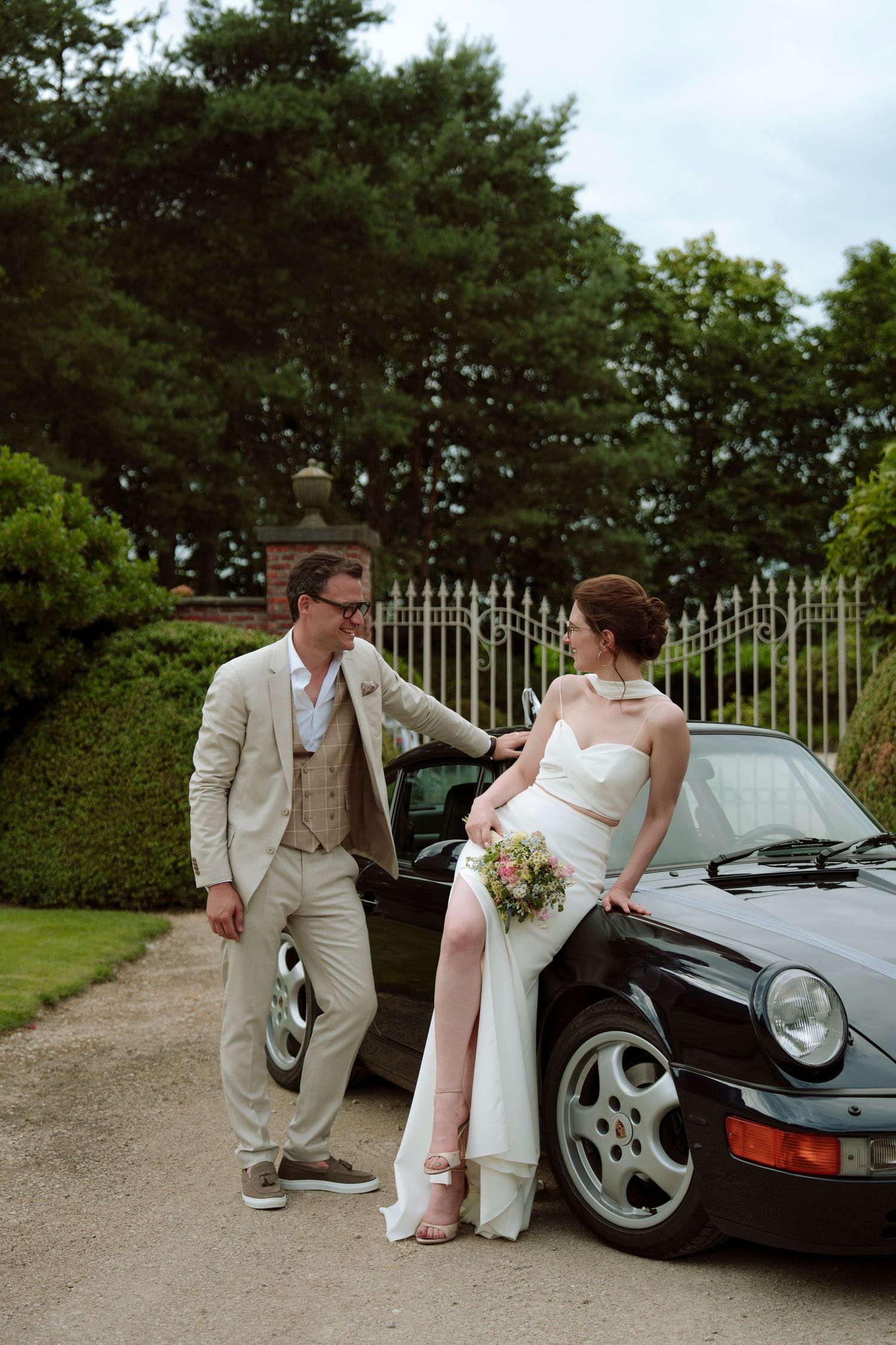 Bride in white gown with slit and groom in beige suit posing beside a classic black Porsche in a garden setting.