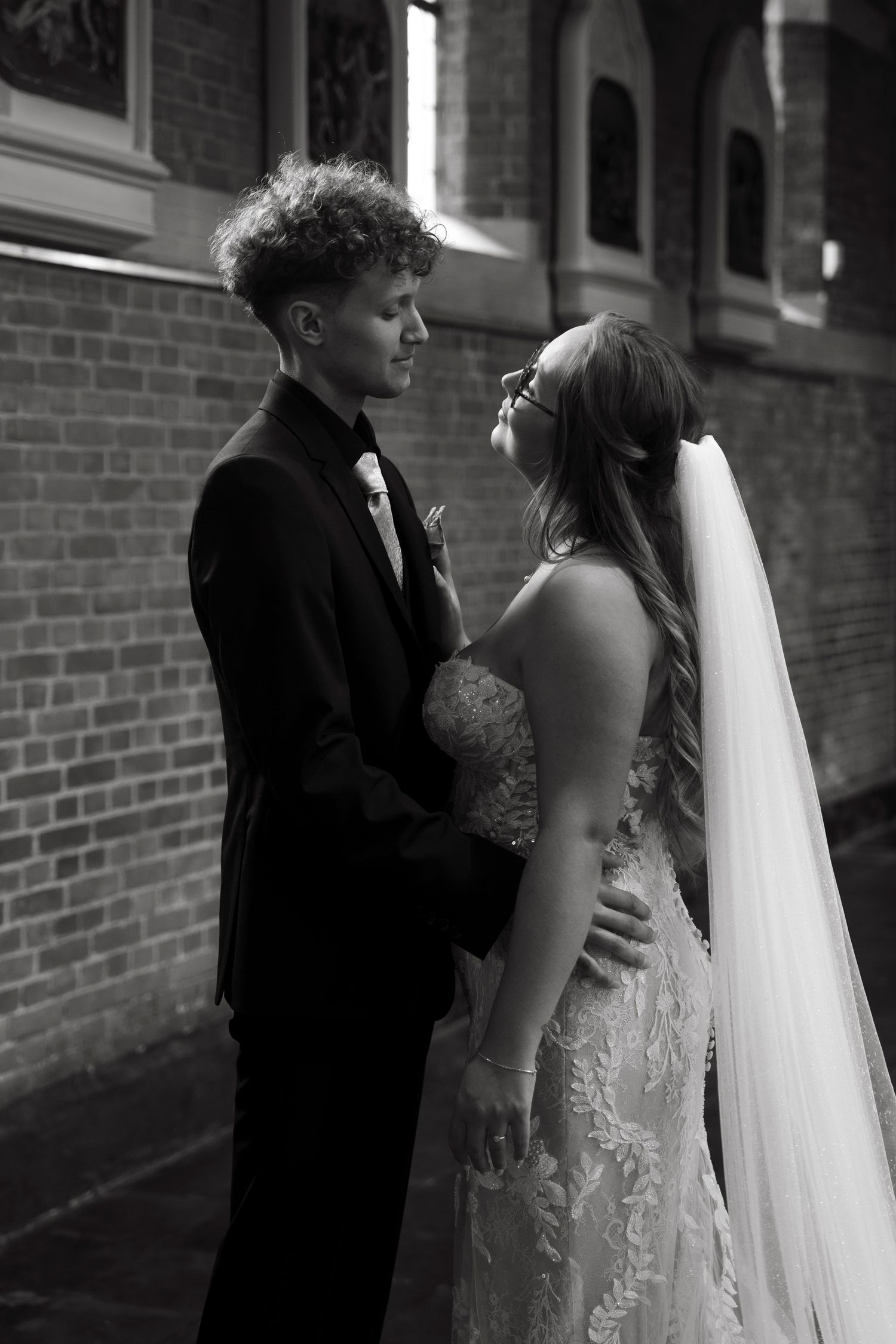 Black and white photo of a bride and groom gazing at each other near a brick wall on their wedding day.