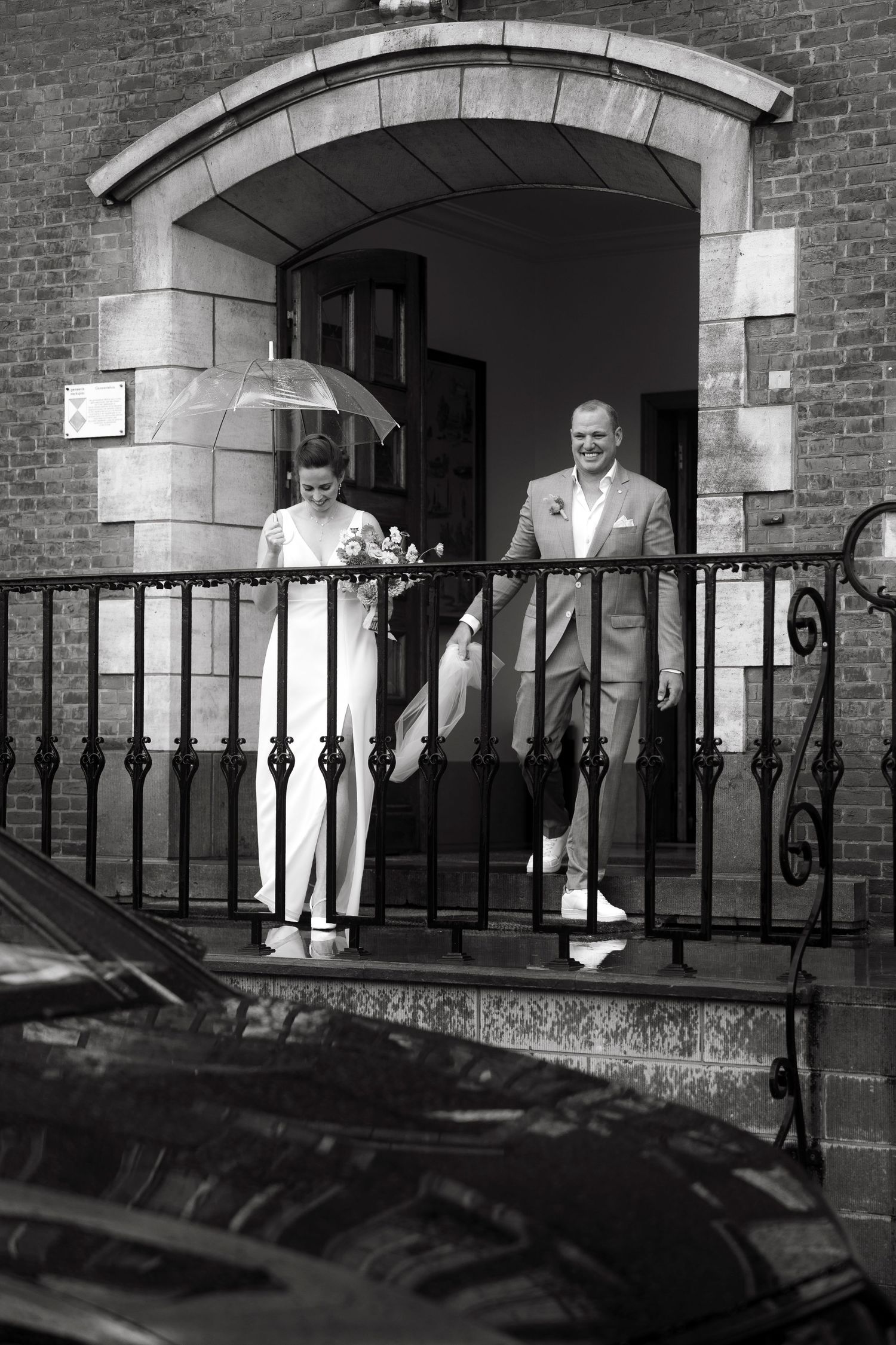 Black and white photo of a bride and groom exiting a brick church, bride holding umbrella and bouquet.