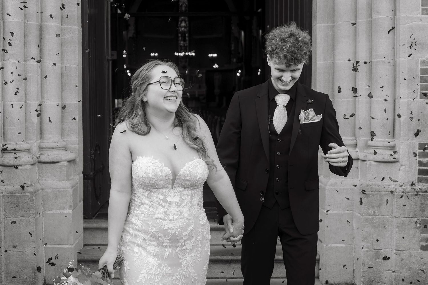 Black and white photo of a smiling bride in lace gown and groom in suit exiting a church with confetti.