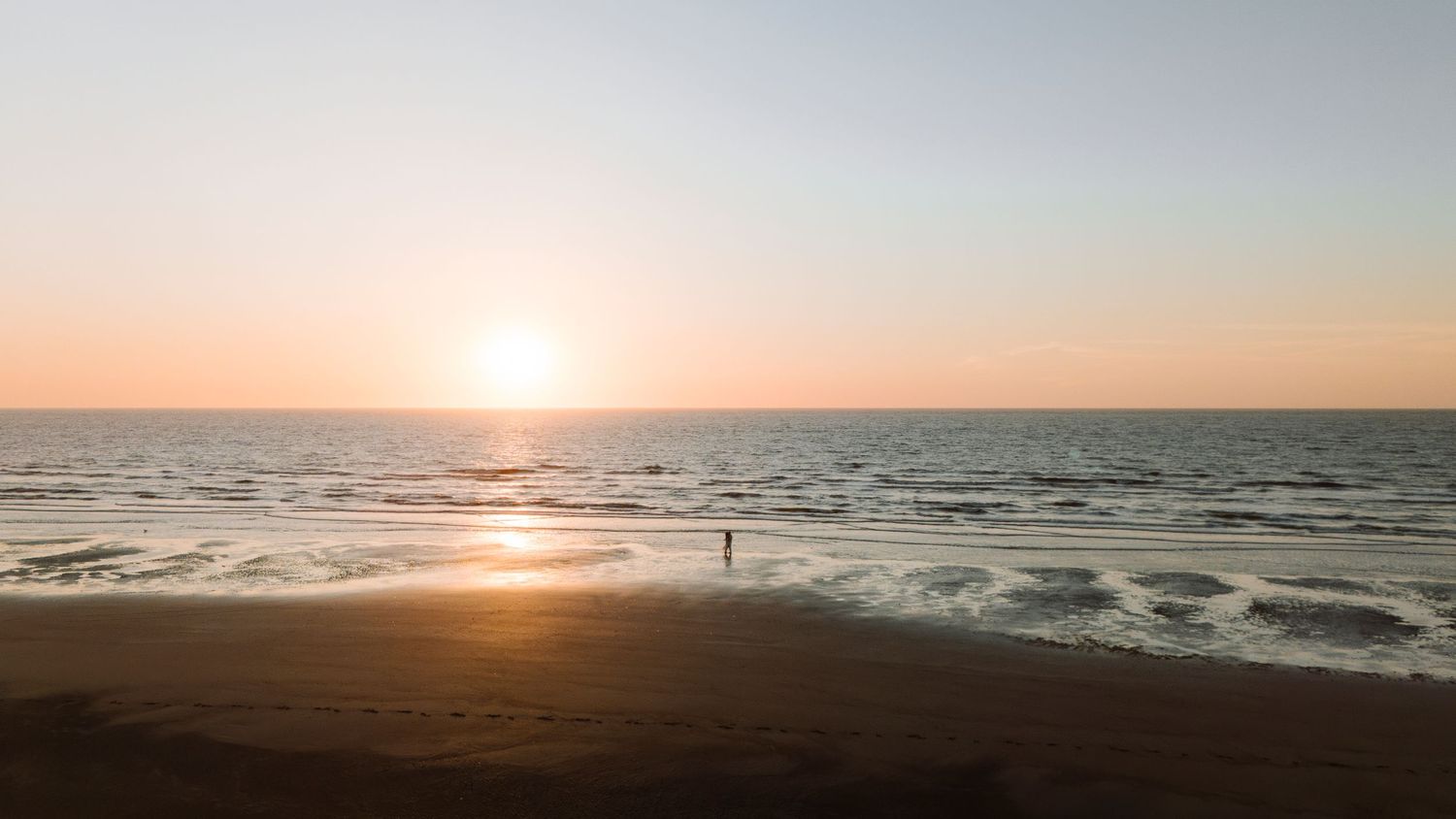 A lone figure stands on a sandy beach at sunset with golden waves and a glowing horizon over the calm ocean.