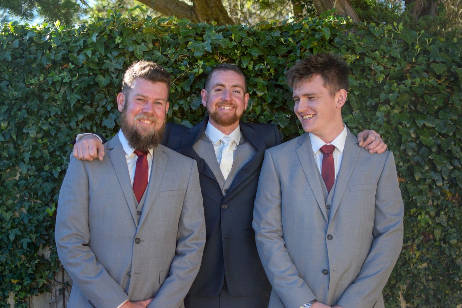 A group in gray suits and burgundy ties pose together for formal wedding party photos outdoors.